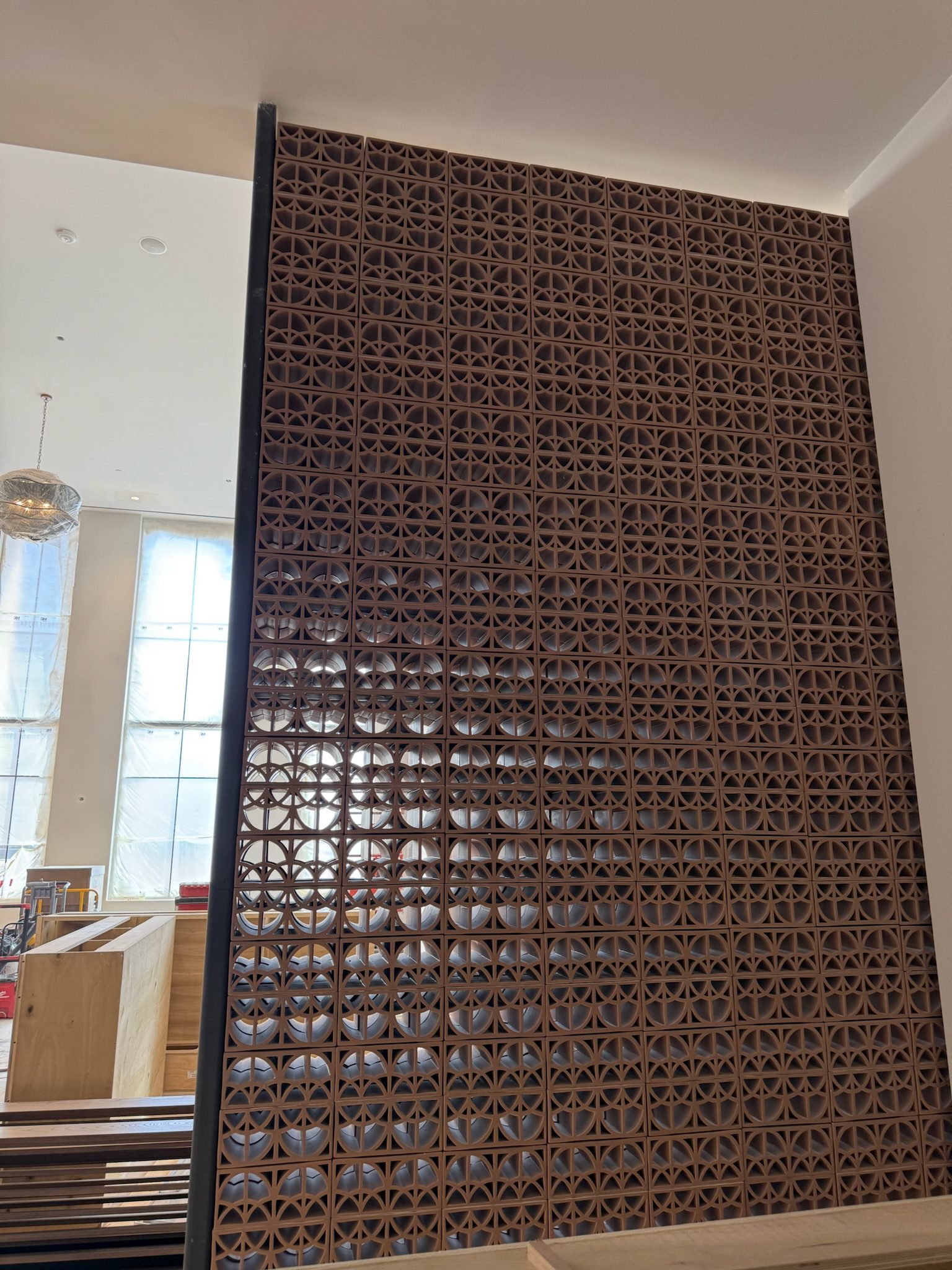 A decorative wall made of brown perforated tiles with a geometric pattern, allowing some light to pass through. The wall is inside a modern, well-lit room with large windows and some wooden furniture nearby.