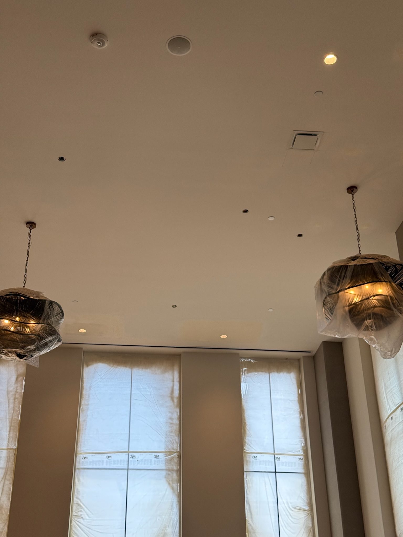A high ceiling with two hanging light fixtures covered in plastic, and tall windows covered with protective sheets, suggesting ongoing renovation or construction in the room.