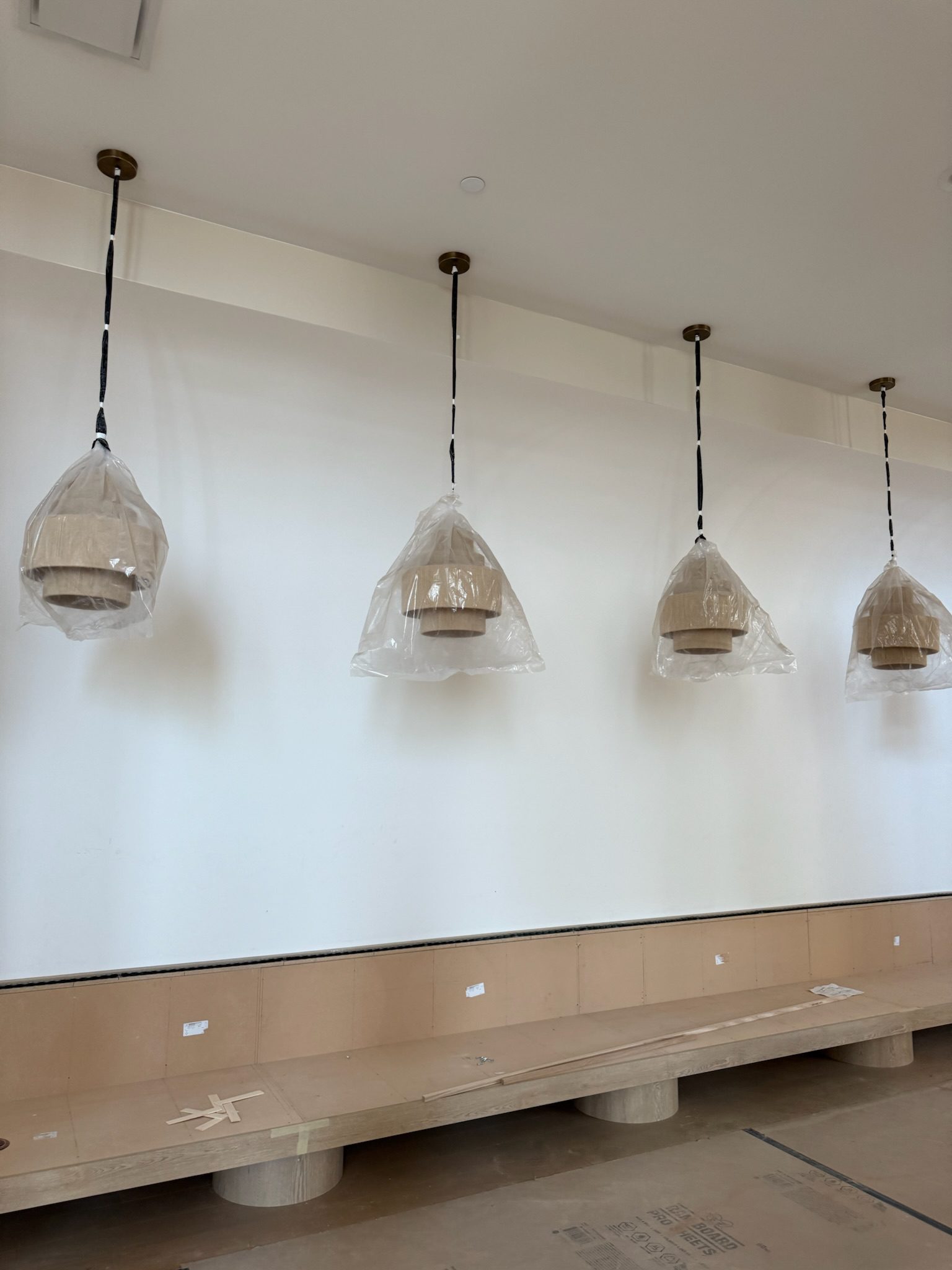 Four pendant lights wrapped in protective plastic hang from the ceiling above a long, unfinished bench in a room under construction with bare walls and floors.