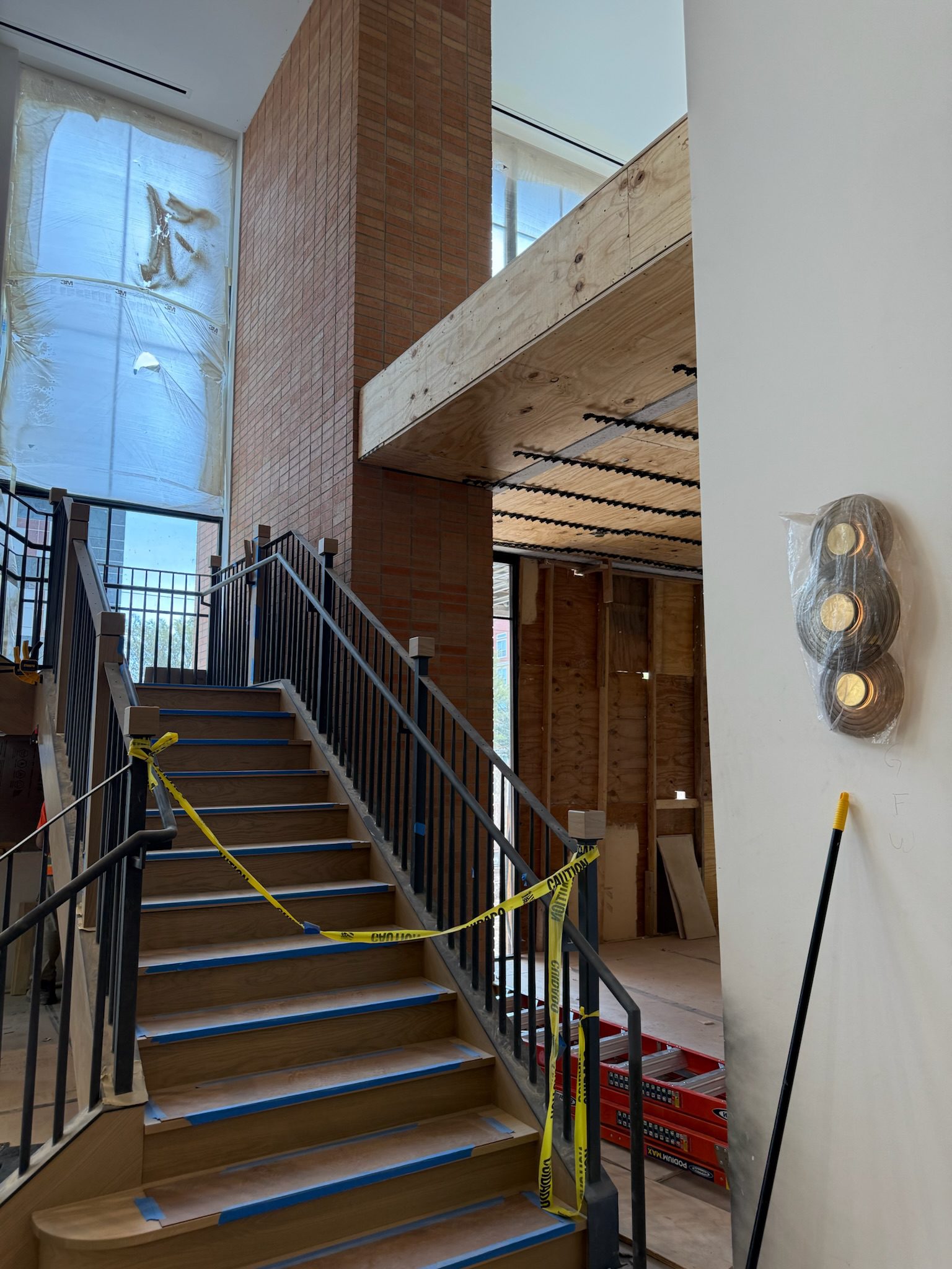 A staircase with black railings and caution tape leads up to a partially constructed loft area in a building under renovation. Walls are unfinished, and a light fixture is wrapped in plastic on the right.