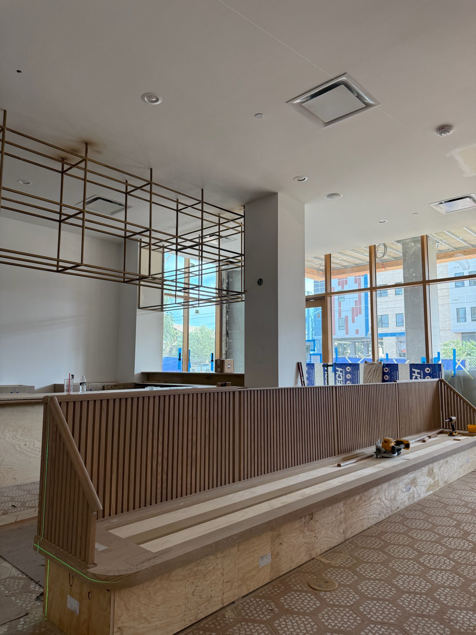 A modern, unfinished interior space with wooden paneling, construction tools on the counter, geometric tile flooring, and large windows letting in natural light. Blue construction barriers are visible outside.