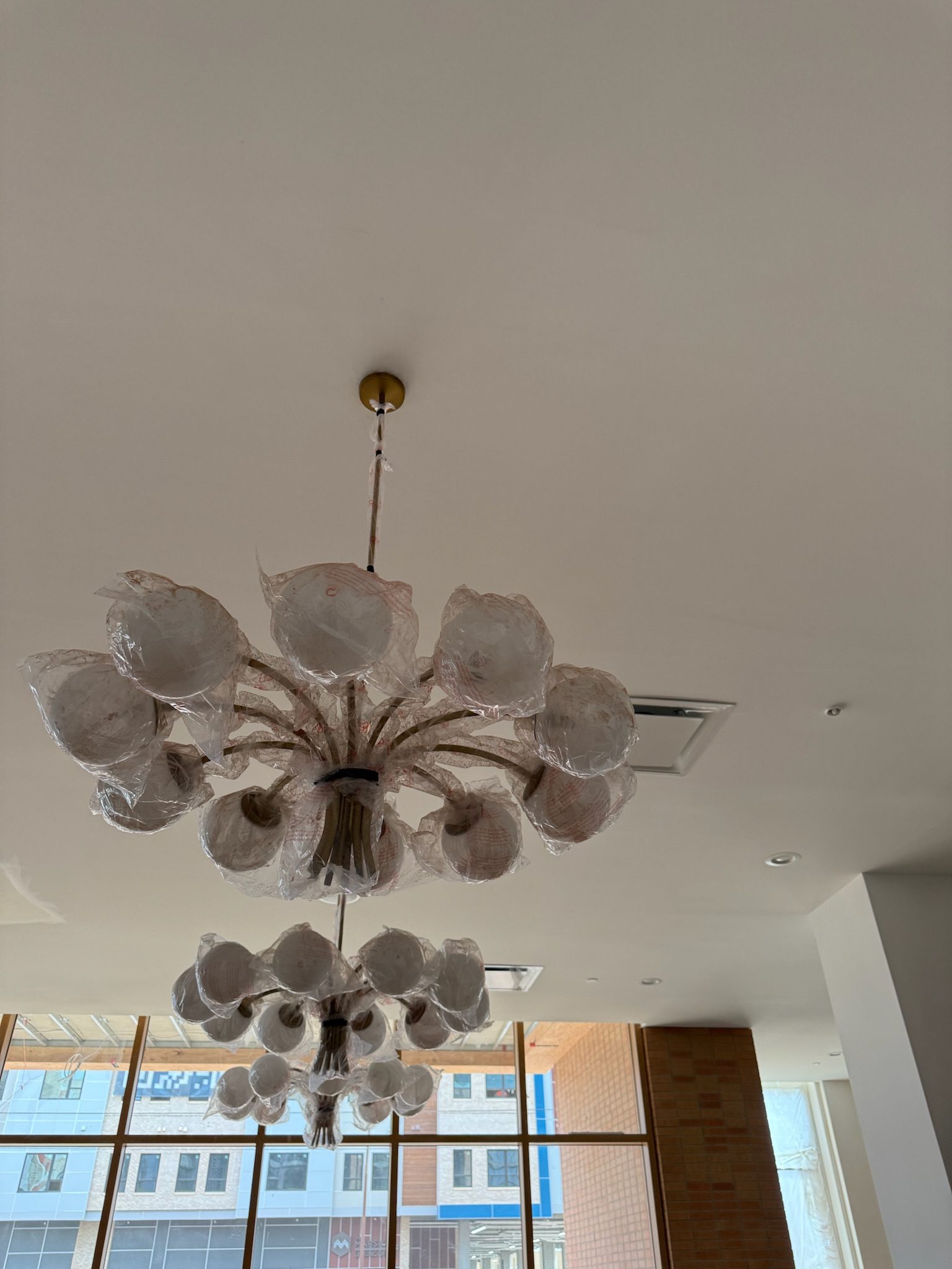 A decorative chandelier wrapped in protective plastic hangs from the ceiling of a modern room with large windows and another identical chandelier in the background.