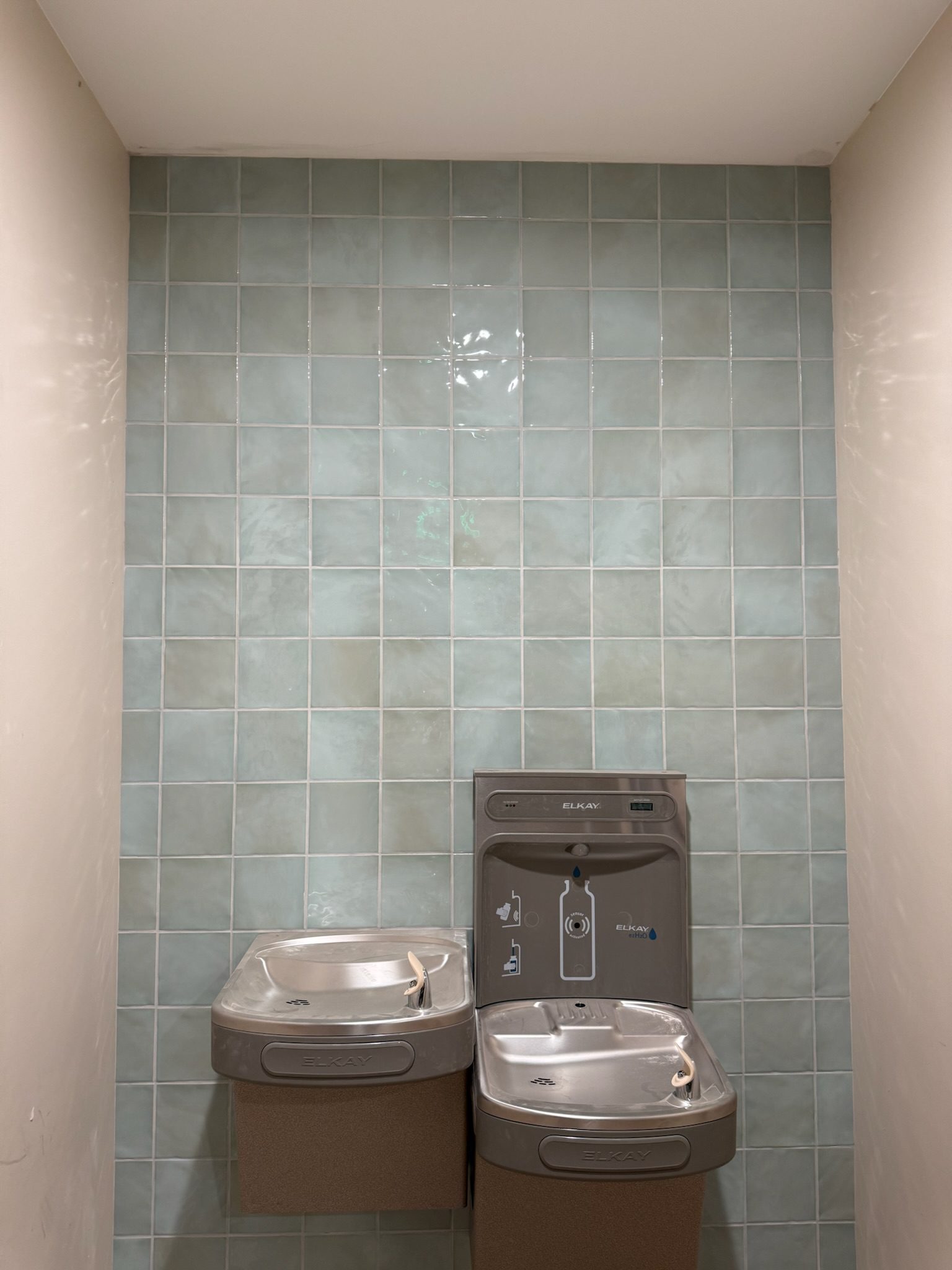 Two drinking fountains and a bottle filling station are mounted on a tiled wall with light greenish-blue square tiles. The fountains are side by side in a small alcove with beige walls.