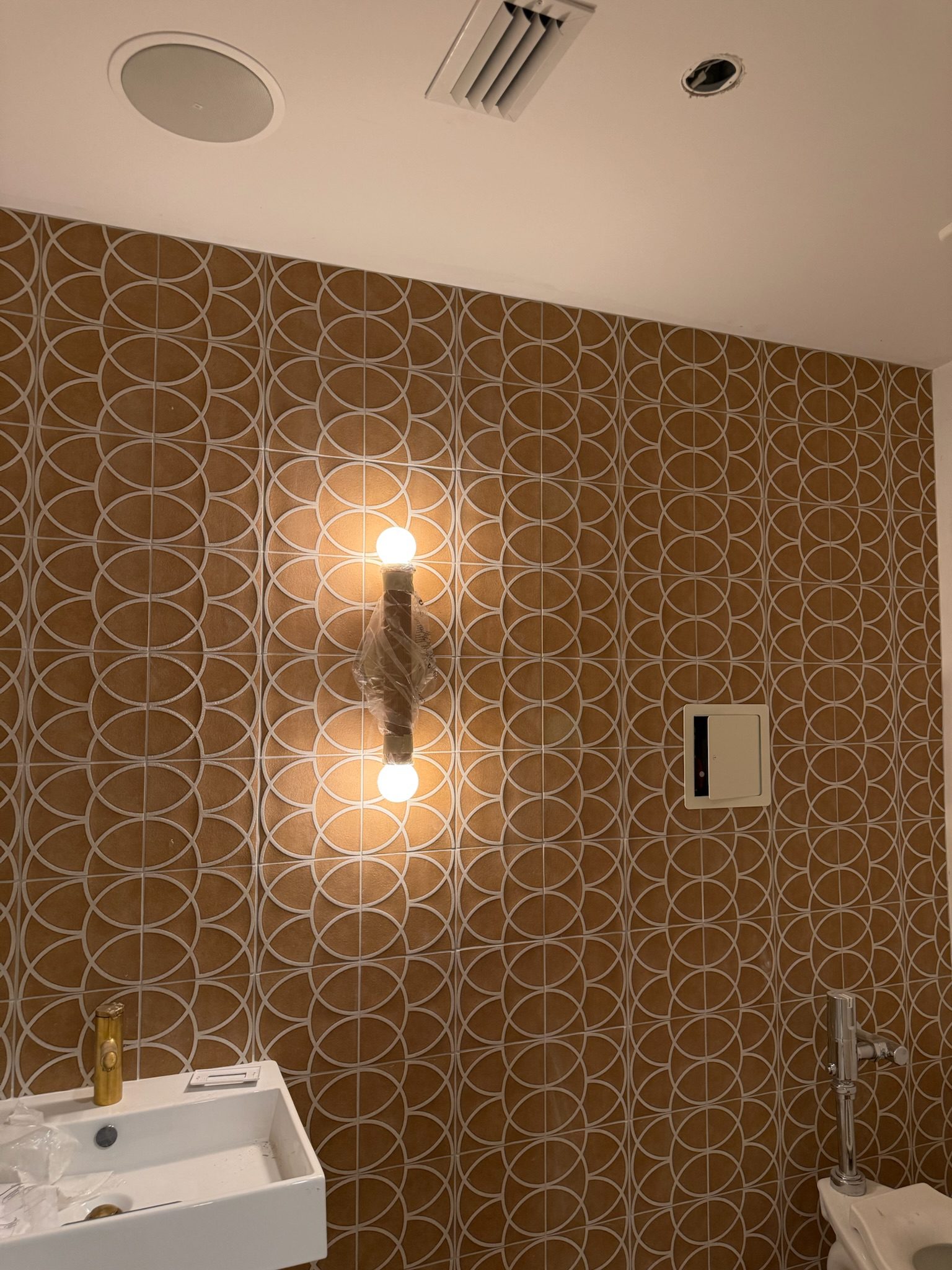 A bathroom wall with brown and white circular patterned wallpaper, a modern light fixture with two exposed bulbs, a small white sink with a gold faucet, and a toilet partially visible on the right.