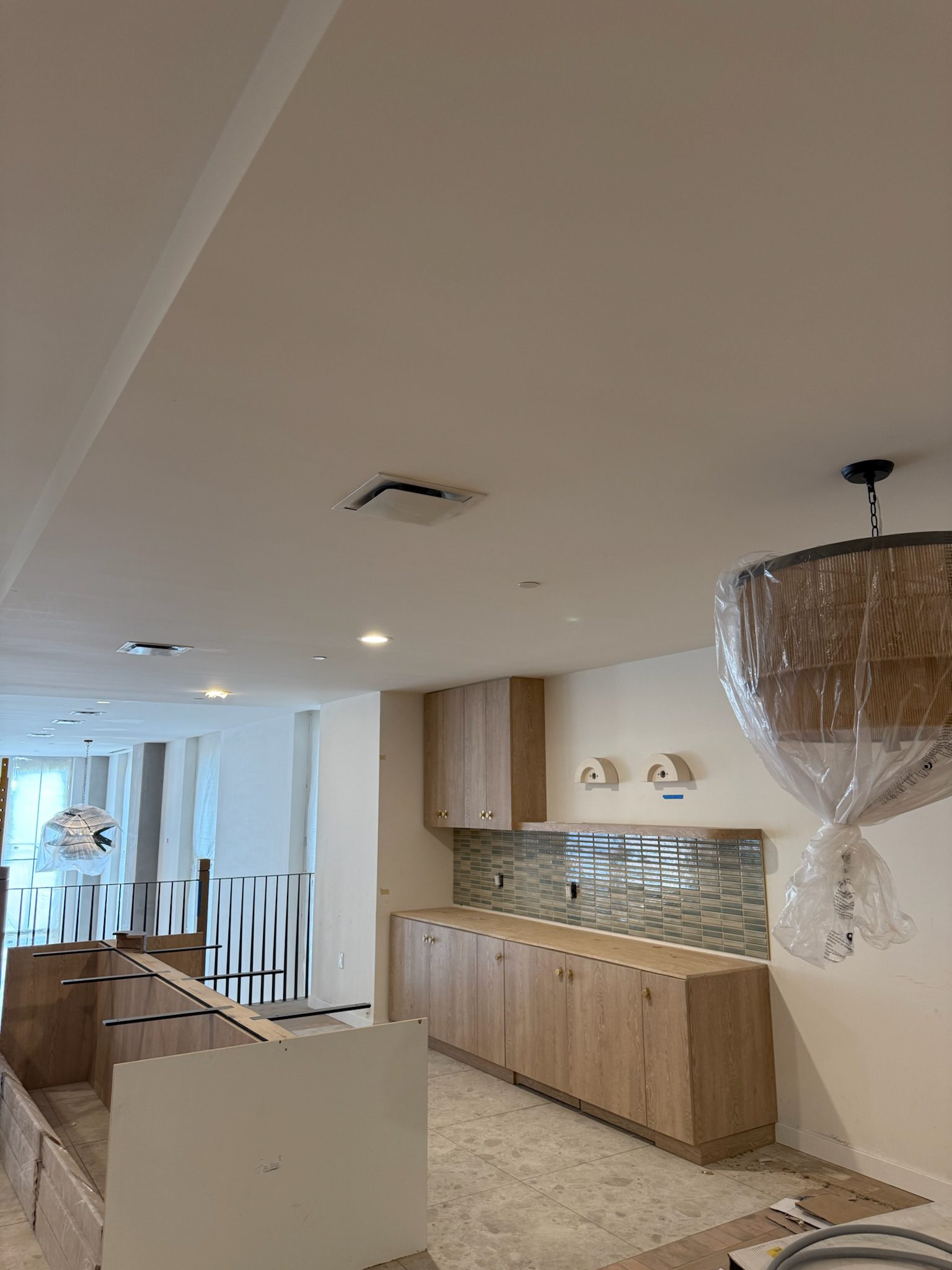 A kitchen under construction with light wood cabinets, a green tile backsplash, unfinished countertops, two wall lights, and pendant lights wrapped in plastic. The floor is unfinished and tools are visible.