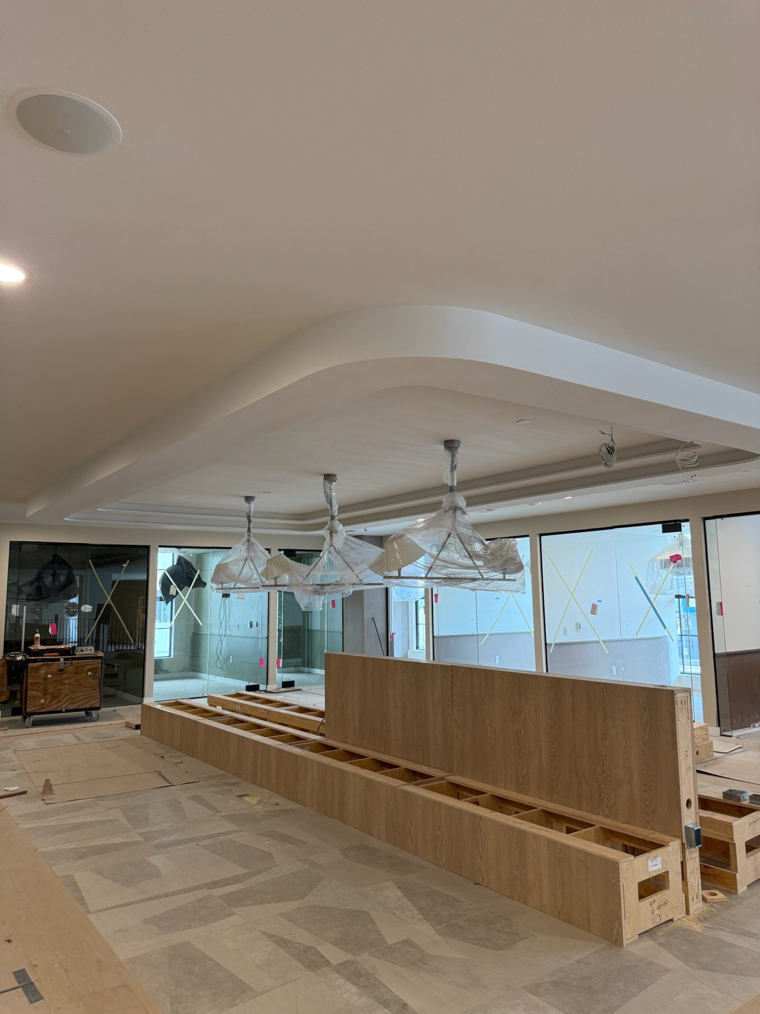 A modern indoor space under construction with a long wooden counter, pendant lights wrapped in plastic, unfinished flooring, and large glass windows and doors in the background.
