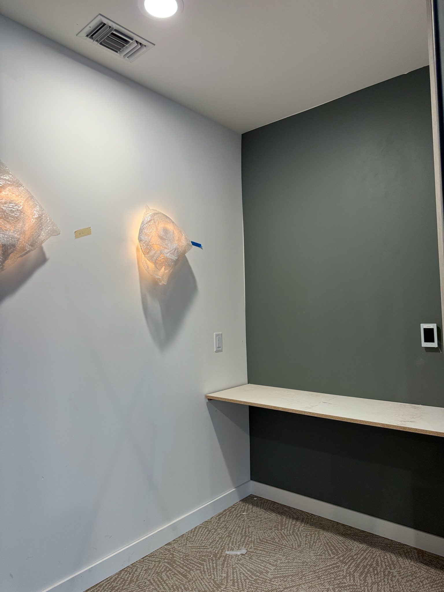 A small room with a dark green accent wall, a built-in floating desk, white walls, carpeted floor, and two wall-mounted lights covered in plastic. An air vent is visible on the ceiling.