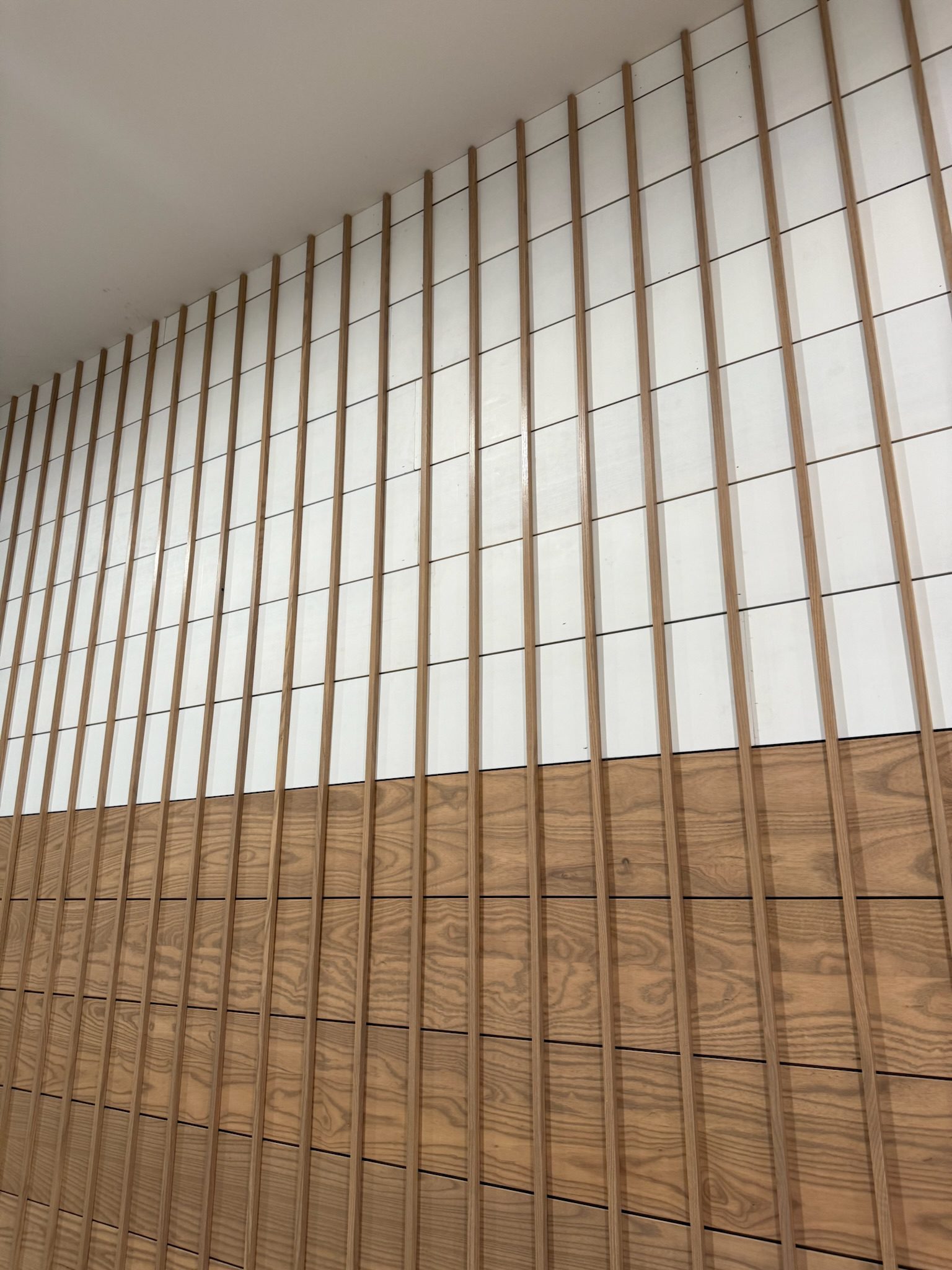 A wall with vertical wooden slats. The lower half features horizontal wooden panels with a natural grain, while the upper half consists of white rectangular panels. The design creates a modern, geometric pattern.