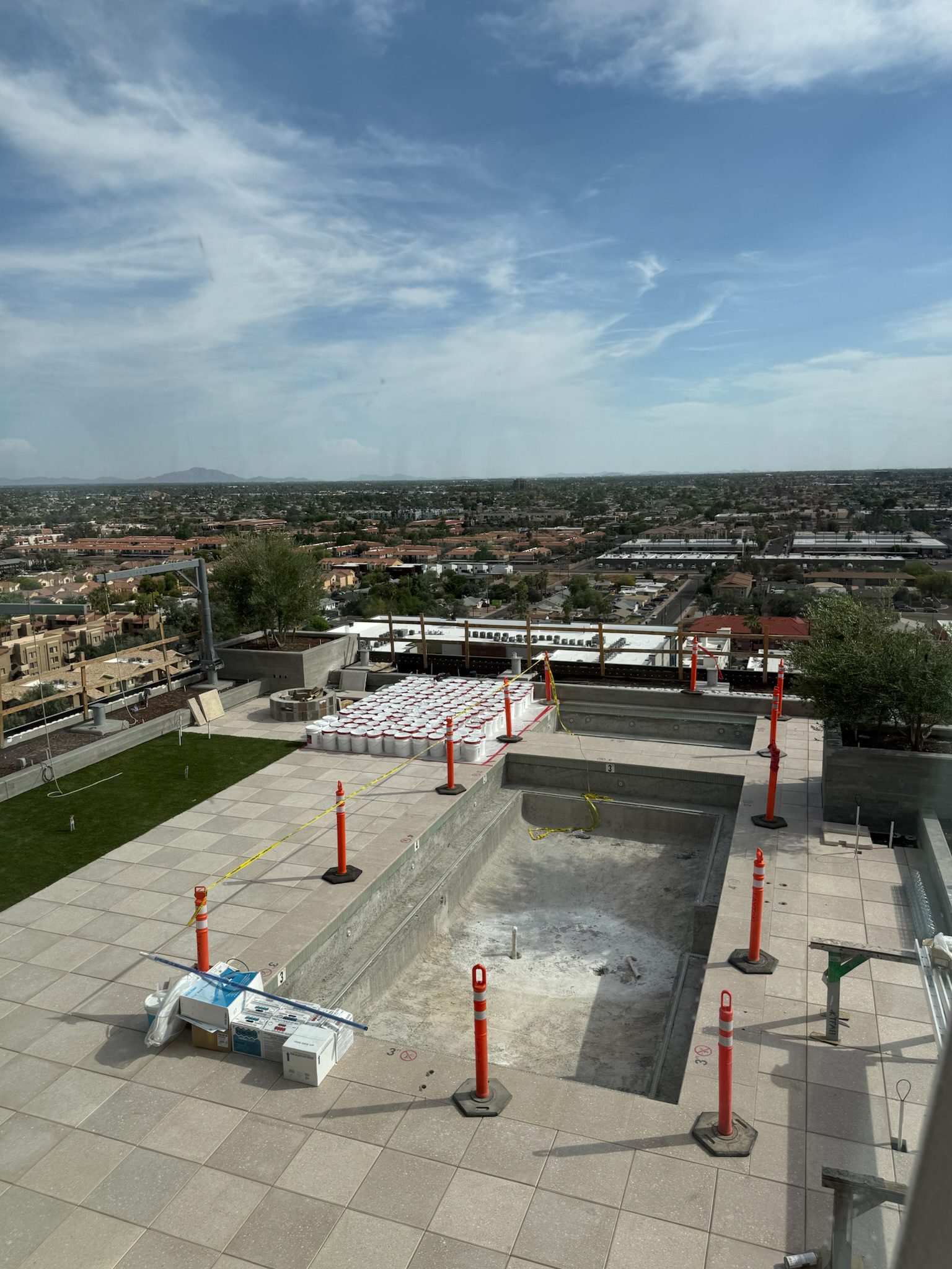 View of an empty rooftop pool under construction, surrounded by orange safety cones and caution tape, with construction materials nearby. In the background, there is a cityscape under a partly cloudy sky.