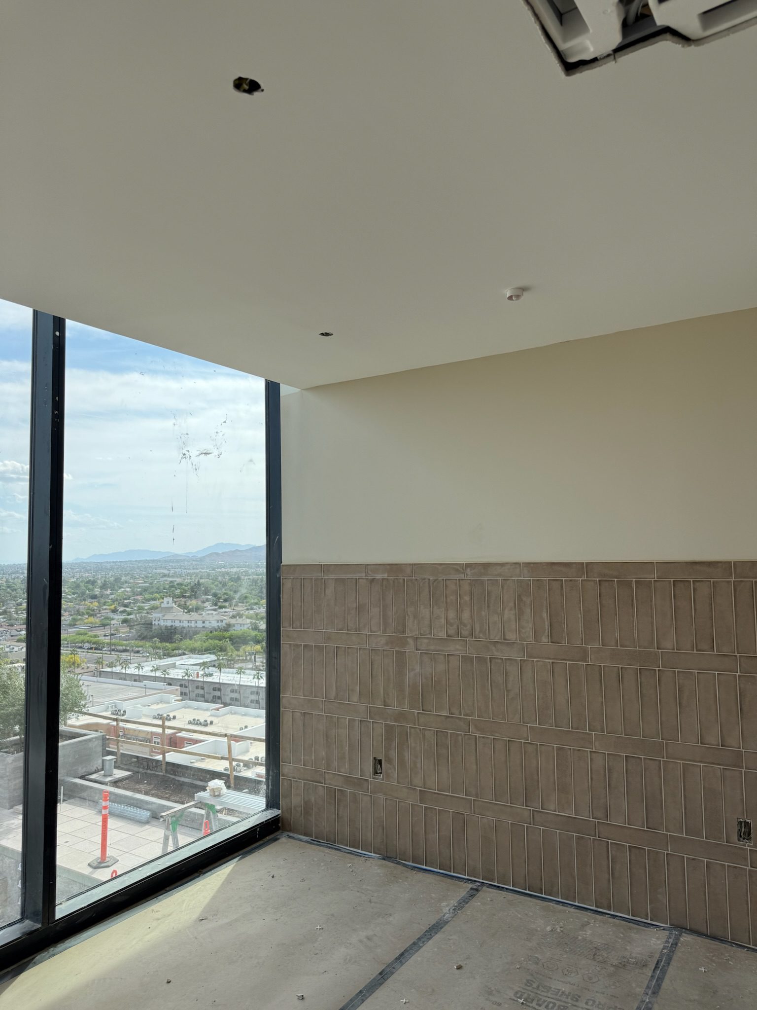 A partially finished room with tan rectangular wall tiles, unpainted drywall, and large floor-to-ceiling windows overlooking a cityscape and mountains under a partly cloudy sky.
