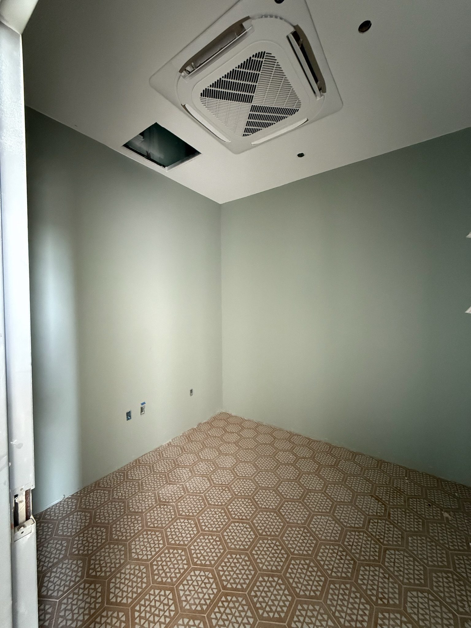 A mostly empty room with pale green walls, patterned beige tile flooring, and a ceiling air conditioning unit. One ceiling tile is missing, revealing an open space above. Light enters from the left side.