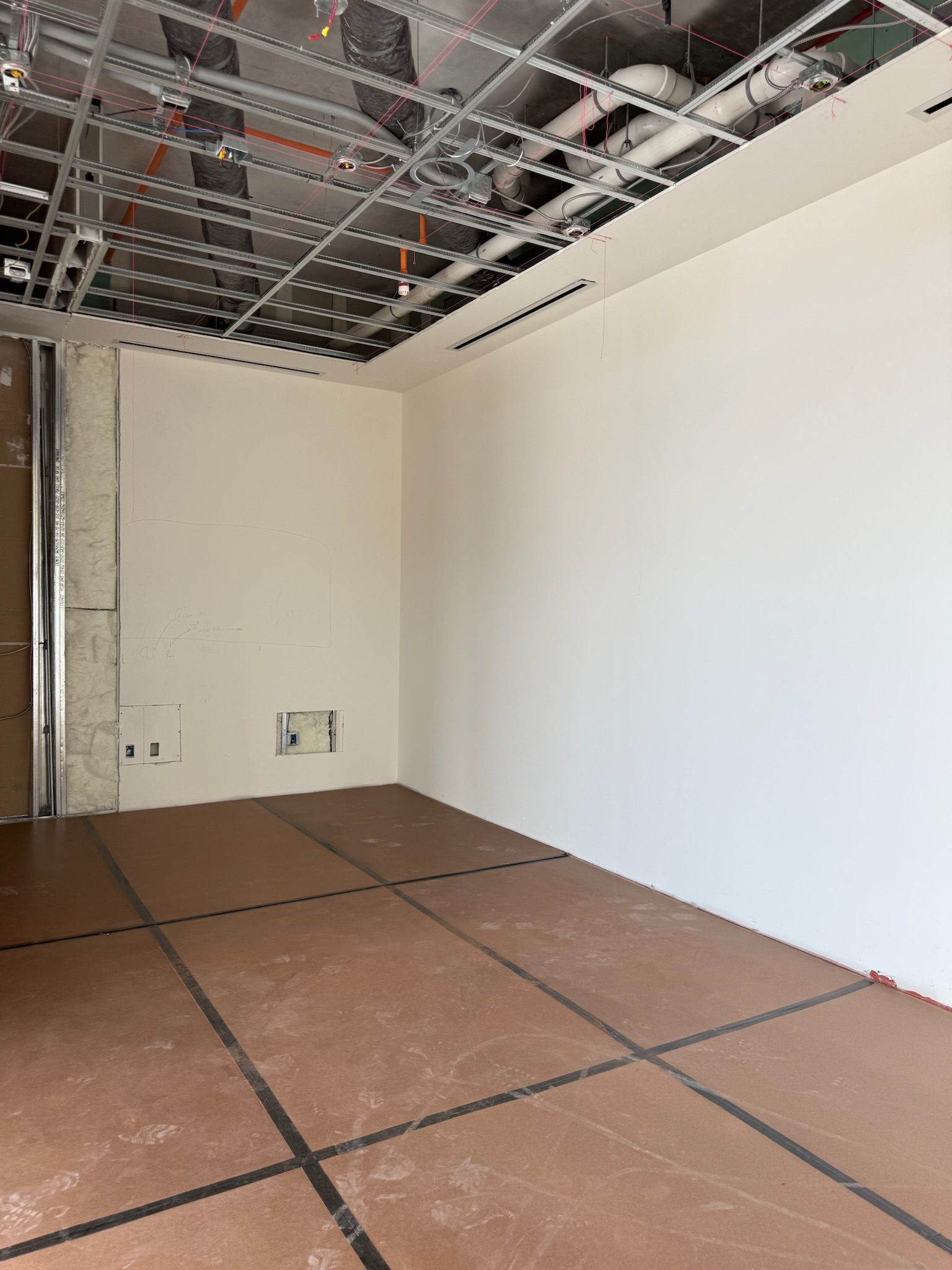 An unfinished room with exposed ceiling pipes and wiring, white walls, and a floor covered with protective brown paper. Some wall panels and electrical outlets are visible.
