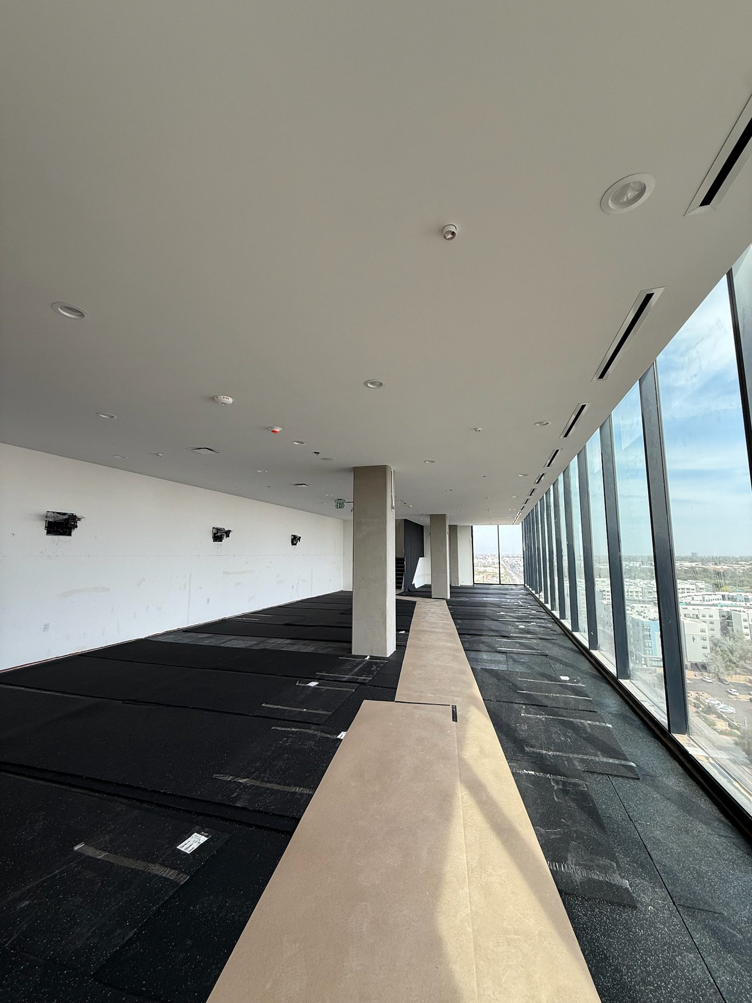 A modern, unfinished high-rise interior with large floor-to-ceiling windows on the right, black flooring, white walls, and exposed columns, overlooking a cityscape under a bright sky.