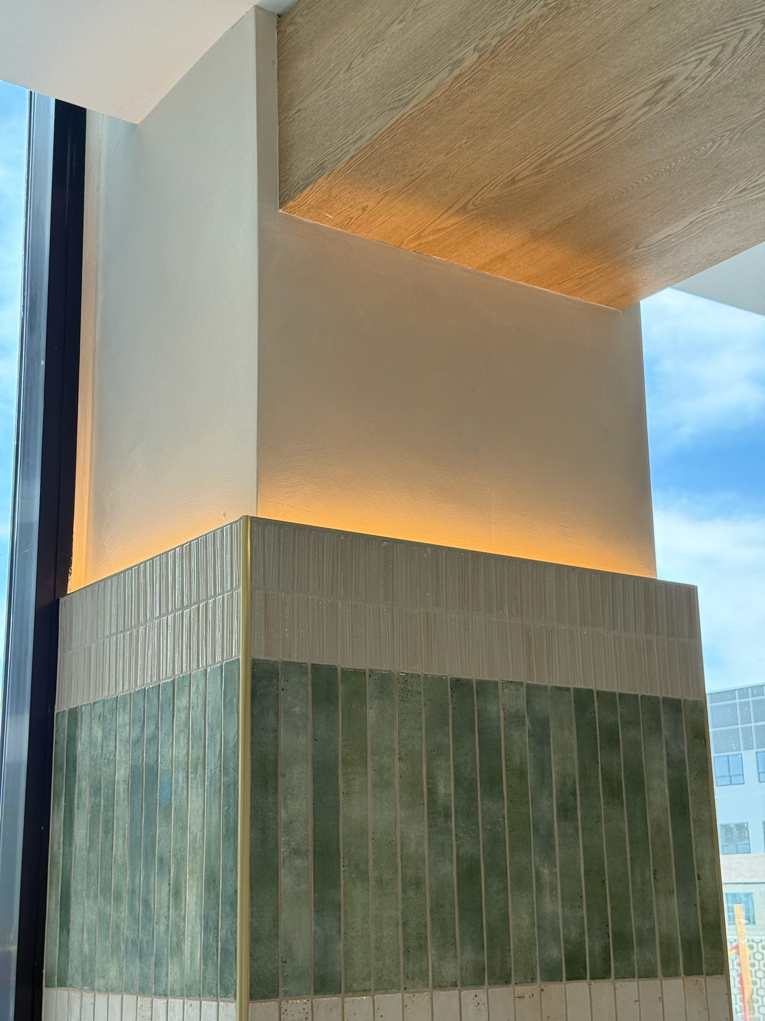 A modern architectural corner with green and cream vertical tiles, a wooden ceiling feature, and soft yellow lighting, next to a large window with a view of blue sky and city buildings outside.