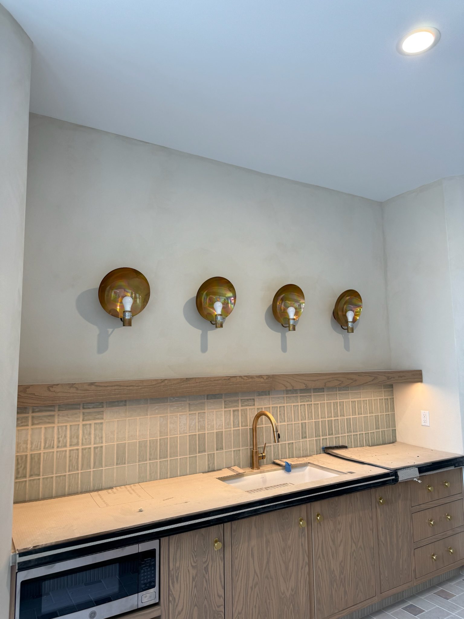 A modern kitchen with light wood cabinets, a white sink, a gold faucet, beige tiled backsplash, and four gold wall sconces above the counter. The counter is partially covered for protection.