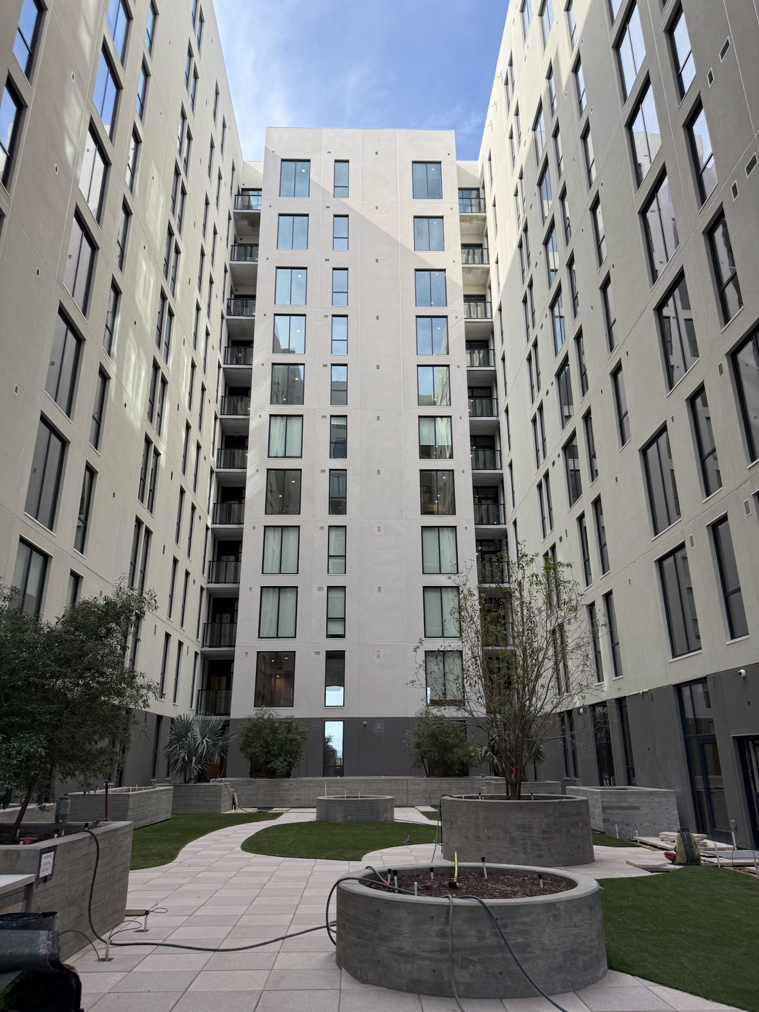 A modern apartment complex courtyard with tall, light-colored buildings on both sides, landscaped with small trees, green grass, paved walkways, and circular planters under a partly cloudy sky.