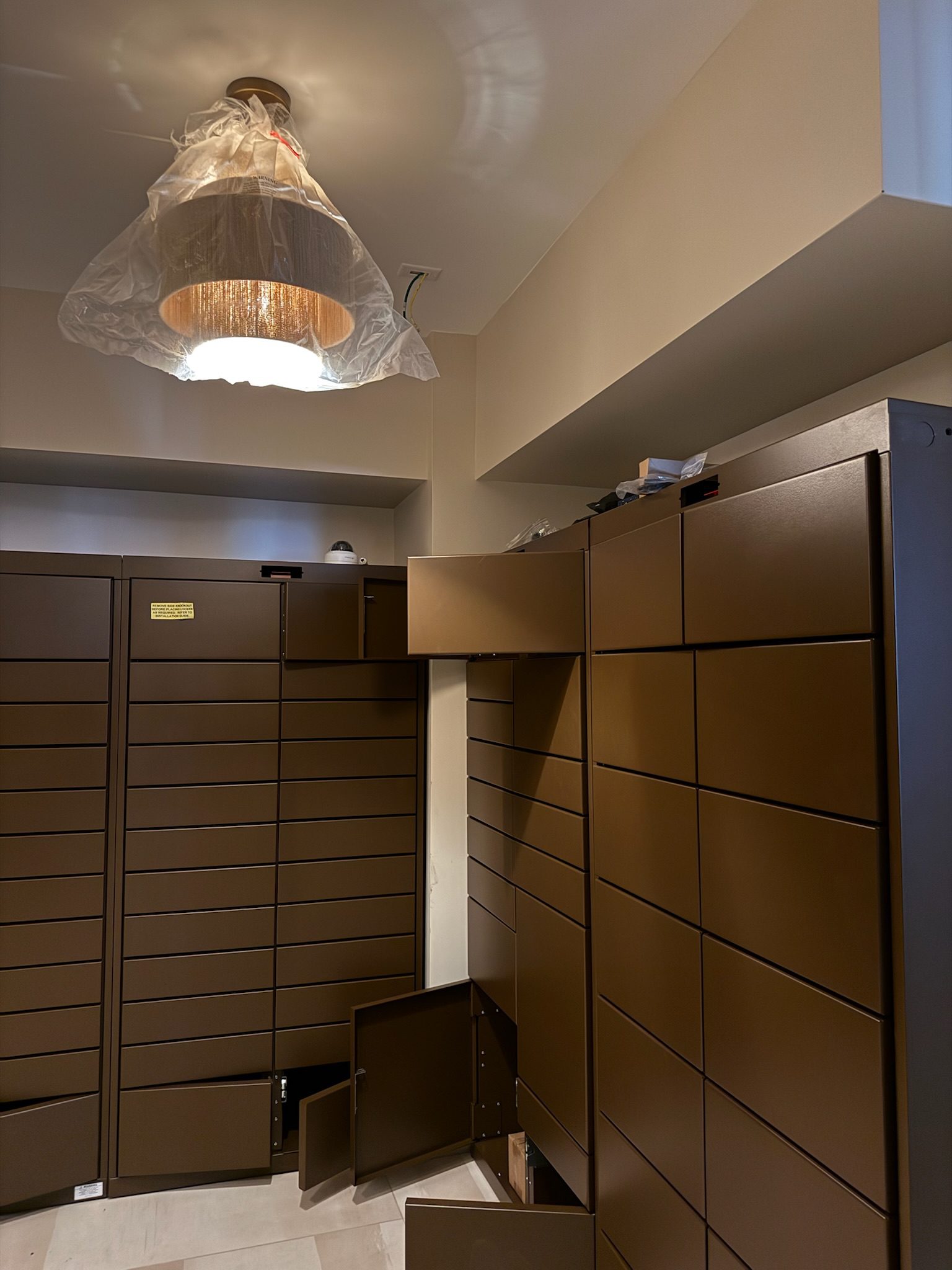 Brown lockers with several open doors and drawers are seen under a ceiling light fixture covered with plastic, inside a beige-walled room. Some tools and items rest on top of the lockers.