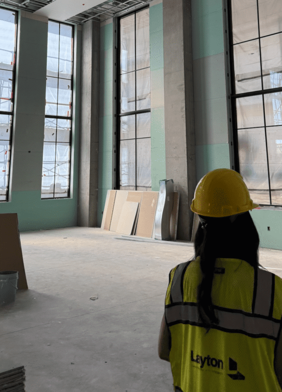 A person in a yellow hard hat and safety vest stands inside a large building during a hard hat tour, surrounded by tall windows, green drywall, exposed ceilings, and scattered construction materials.