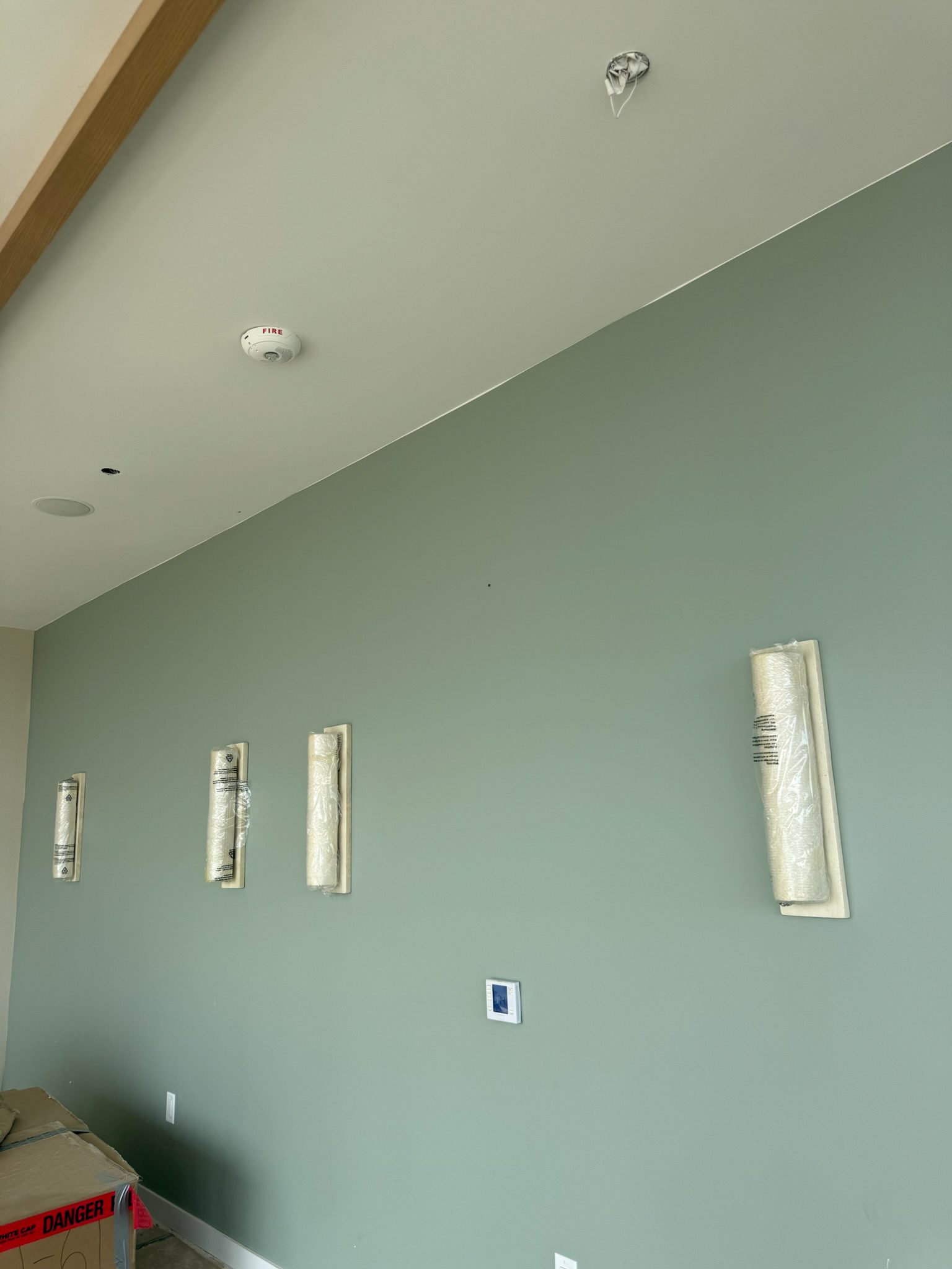 A green wall with four light fixtures covered in protective plastic, a smoke detector on the white ceiling, and a cardboard box partially visible in the bottom left corner. The room appears to be under construction.