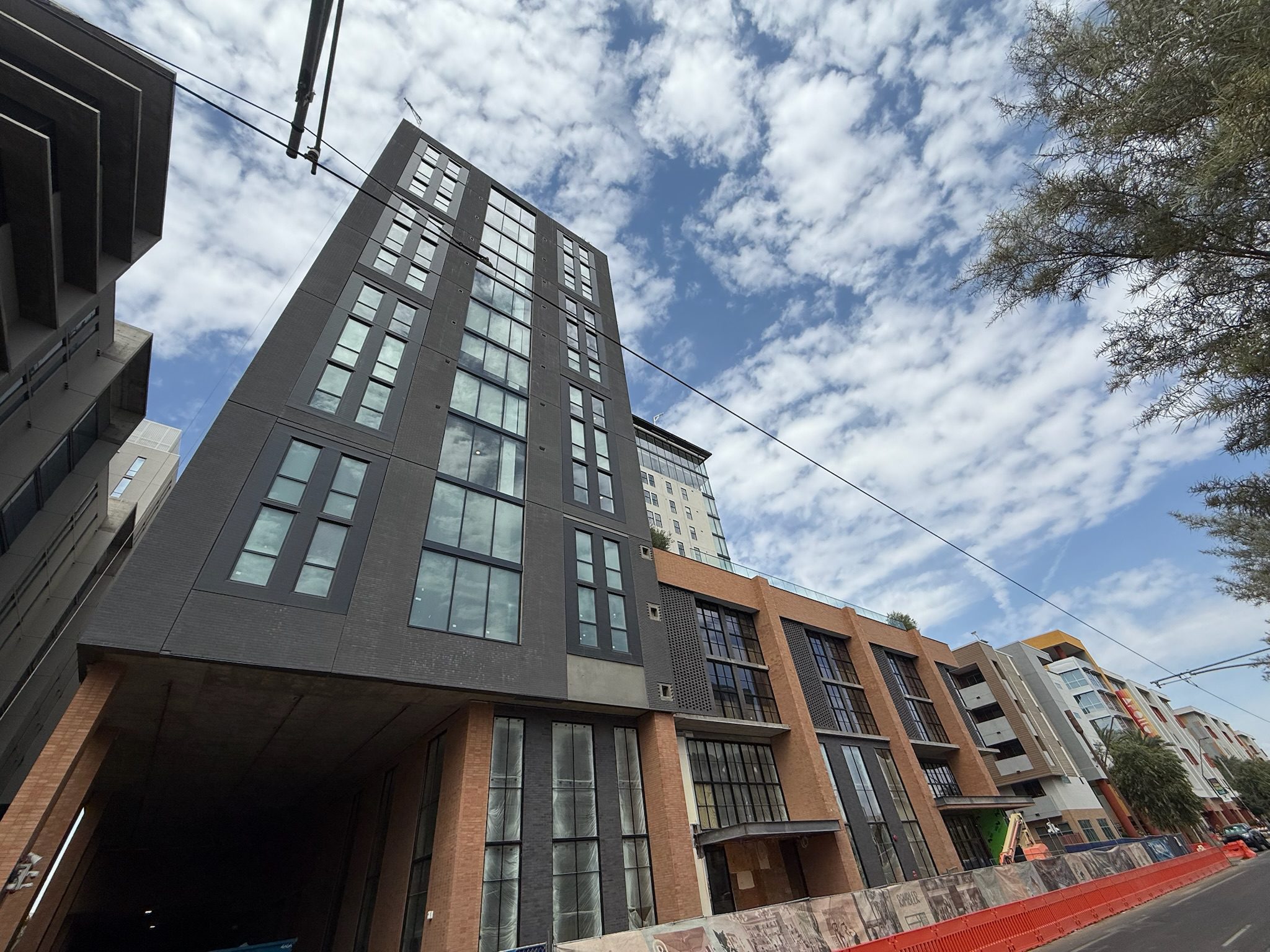 A tall modern building with large windows stands under a partly cloudy sky, surrounded by other urban structures. The street below is lined with red barriers and construction fencing.