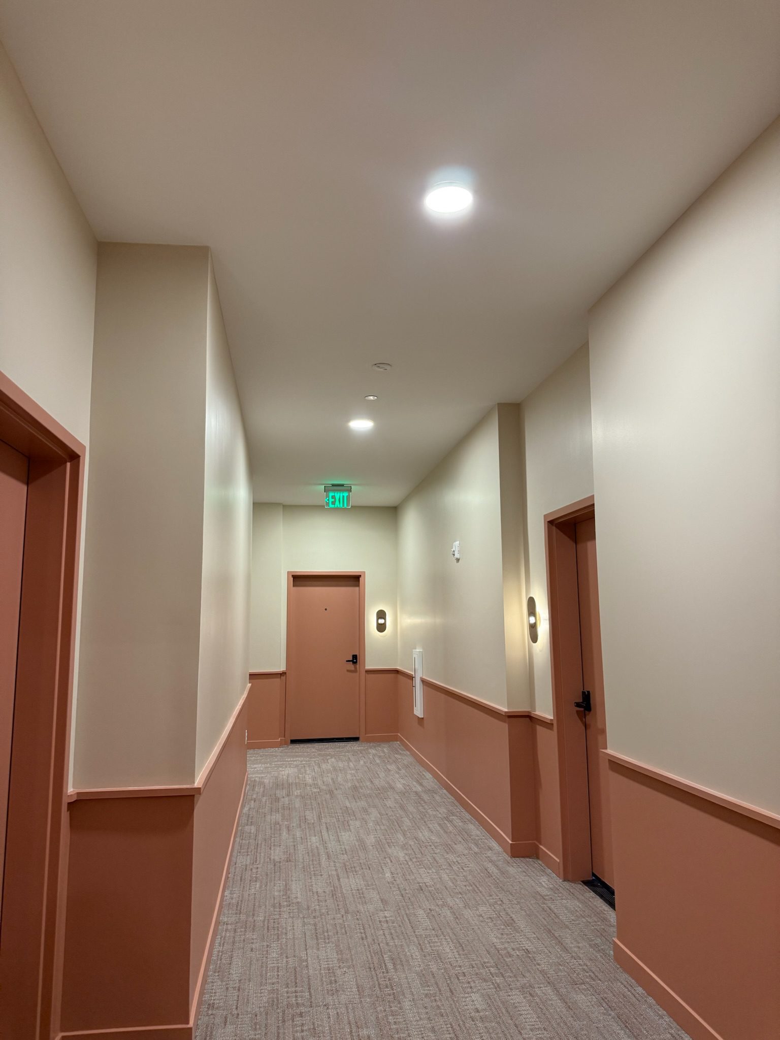 A well-lit hallway in an apartment building with beige walls, salmon-colored doors and trim, carpeted flooring, ceiling lights, and an illuminated green exit sign above the door at the end.