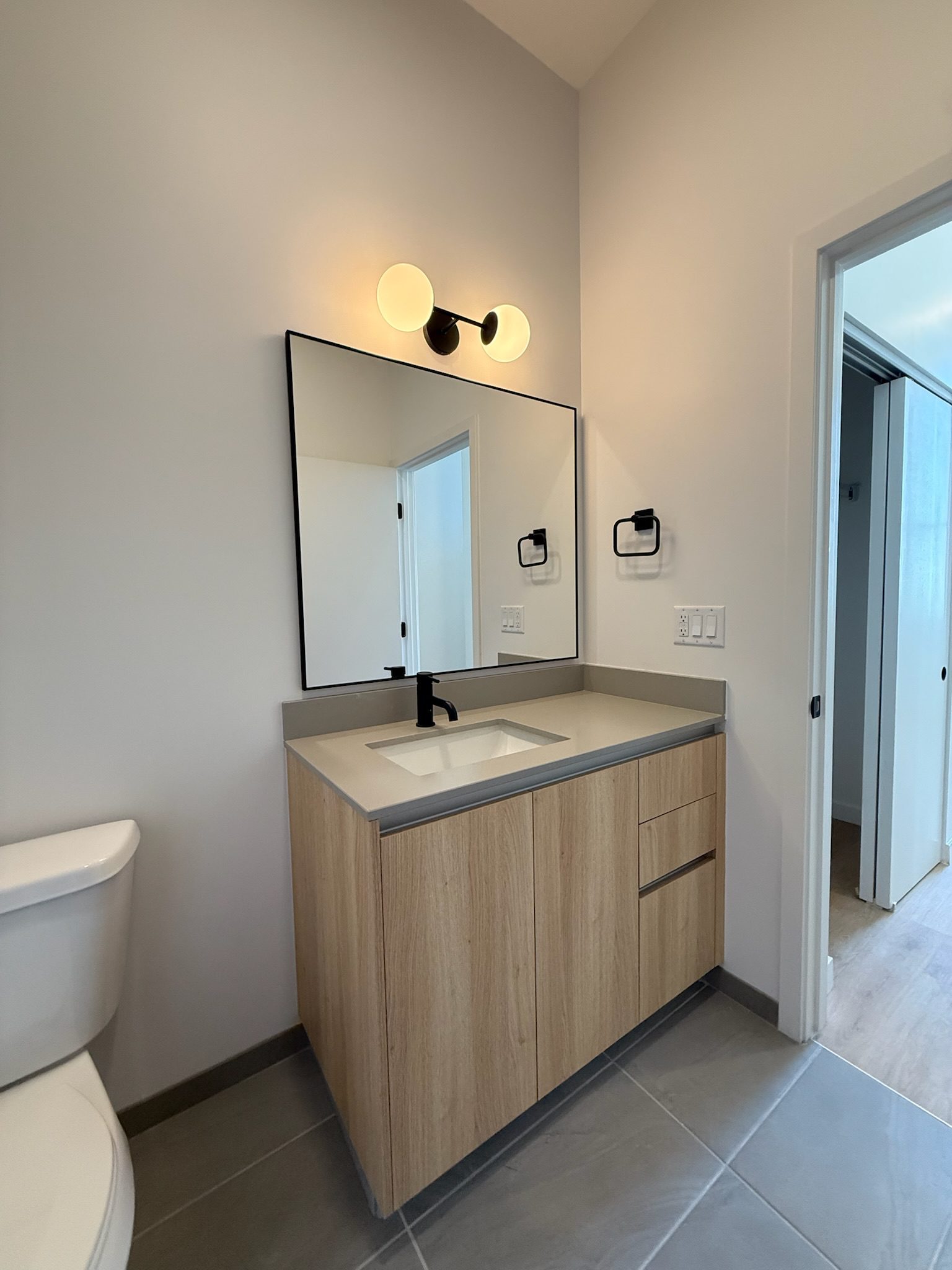 Modern bathroom with a light wood vanity, rectangular mirror, black faucet, wall-mounted light fixture, and gray tile floor. Toilet is partially visible on the left, and doors to other rooms are open.