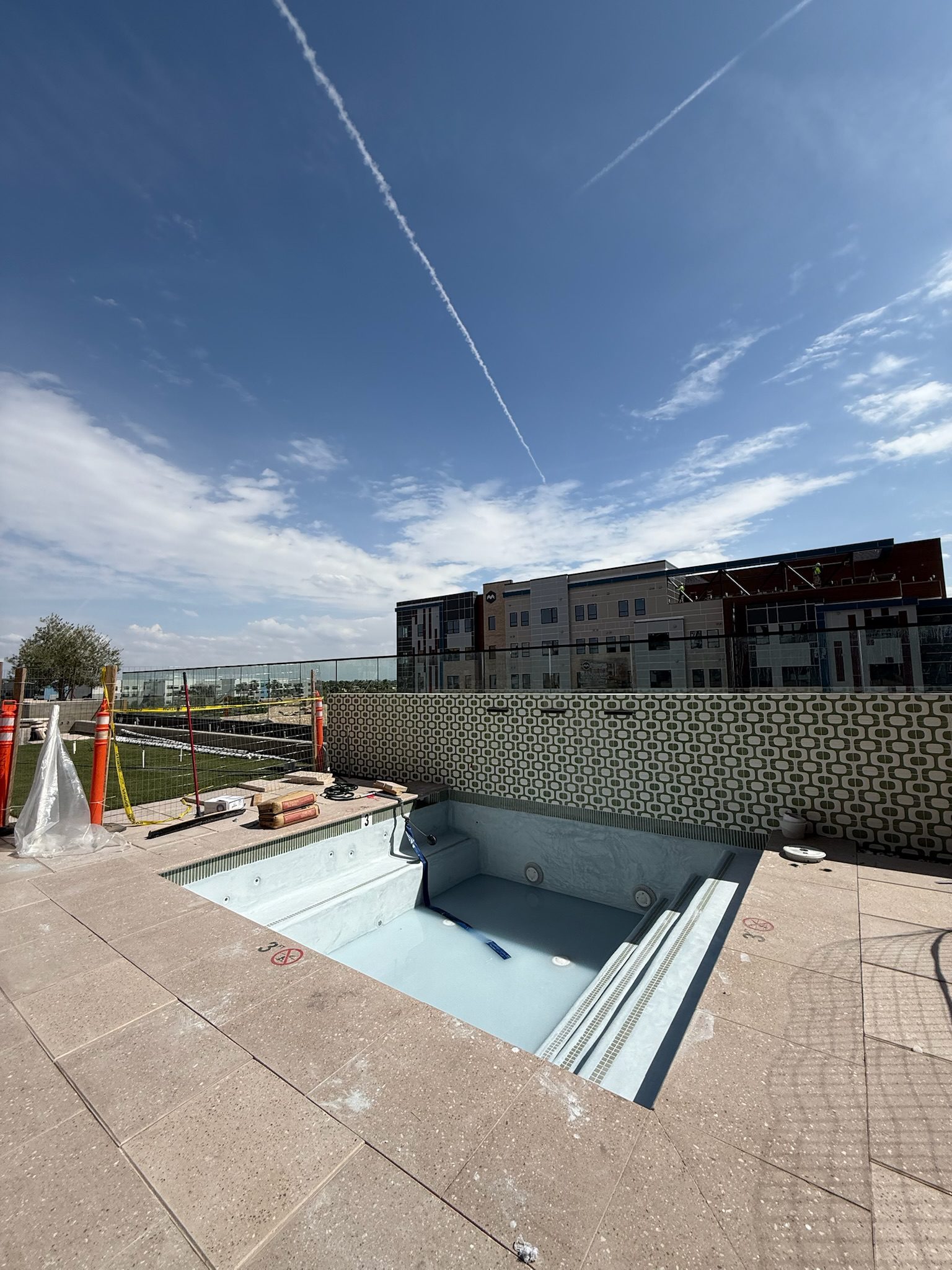An empty swimming pool under construction is surrounded by tiles and building materials, with a patterned wall and a modern apartment building in the background under a blue sky with a visible jet contrail.