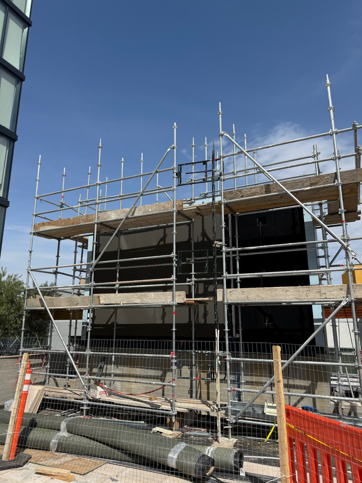A modern building under construction, surrounded by scaffolding and fencing. Construction materials are on the ground, and the sky is clear and blue above.