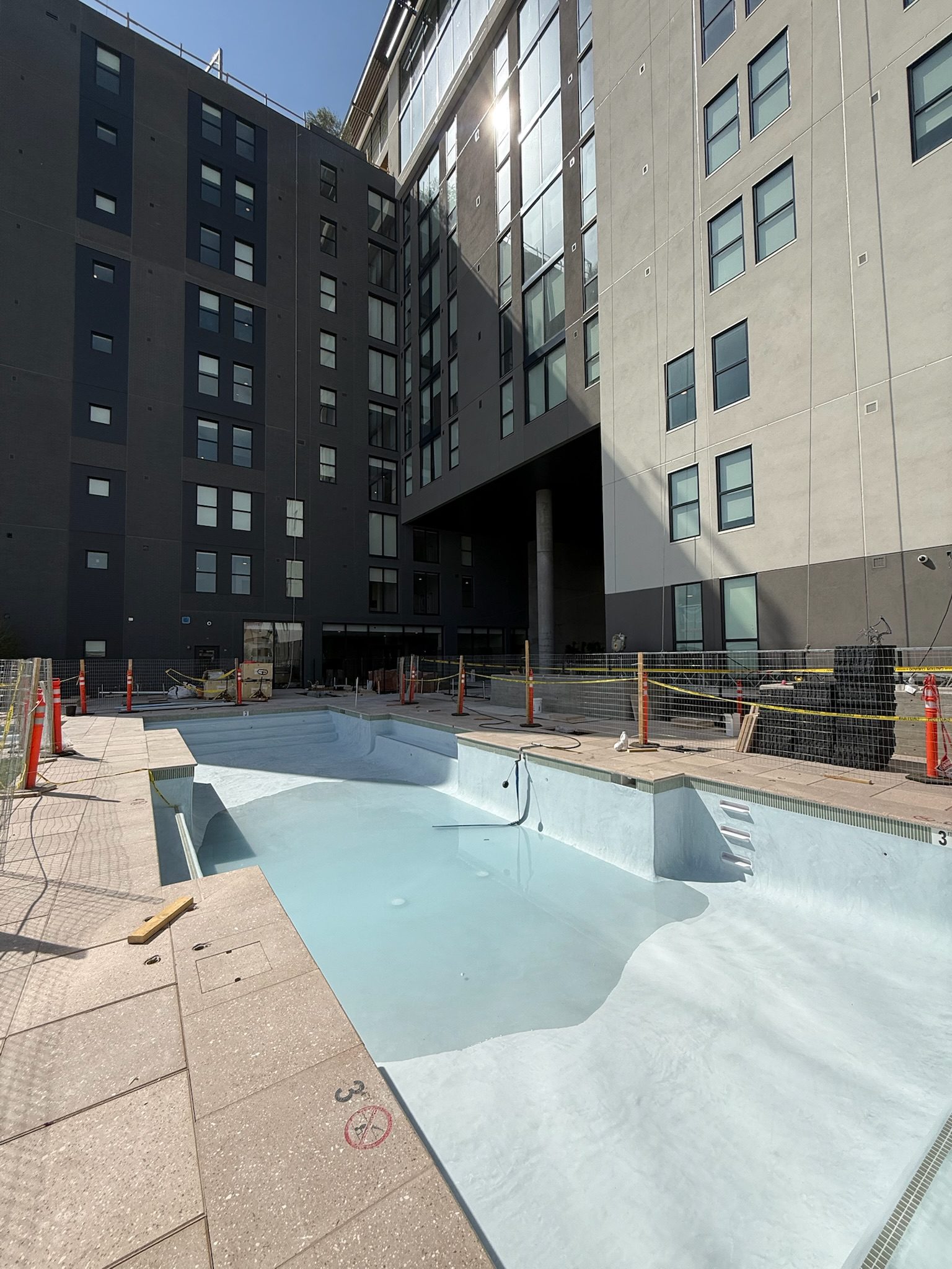 An empty swimming pool with a small amount of water at the bottom is surrounded by a modern apartment building. Construction materials, caution tape, and orange cones are visible around the pool area under bright sunlight.