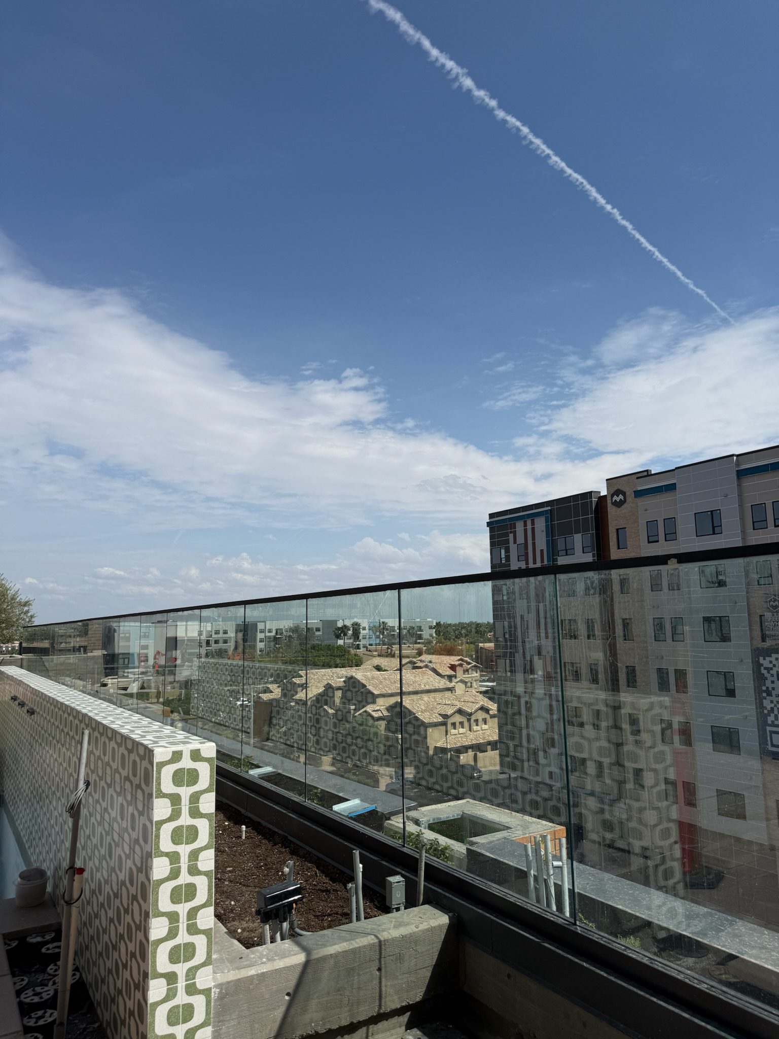View from a balcony with a glass railing, overlooking modern apartment buildings and a house. The sky is mostly clear with a few clouds and a white contrail stretching diagonally overhead.
