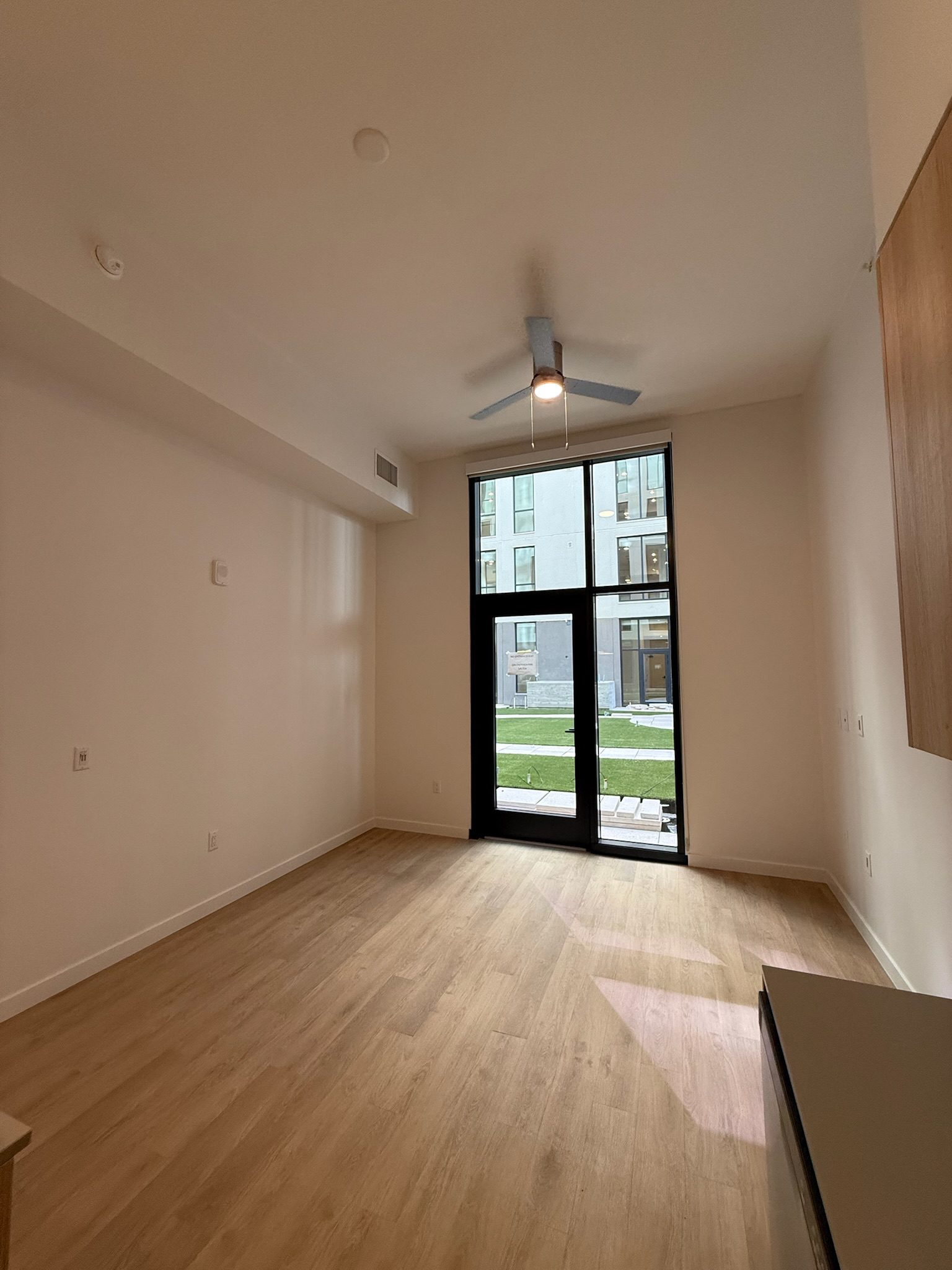 A modern, empty room with light wood flooring, tall ceiling, ceiling fan, and large floor-to-ceiling windows and door facing a courtyard with grass and an apartment building visible outside.
