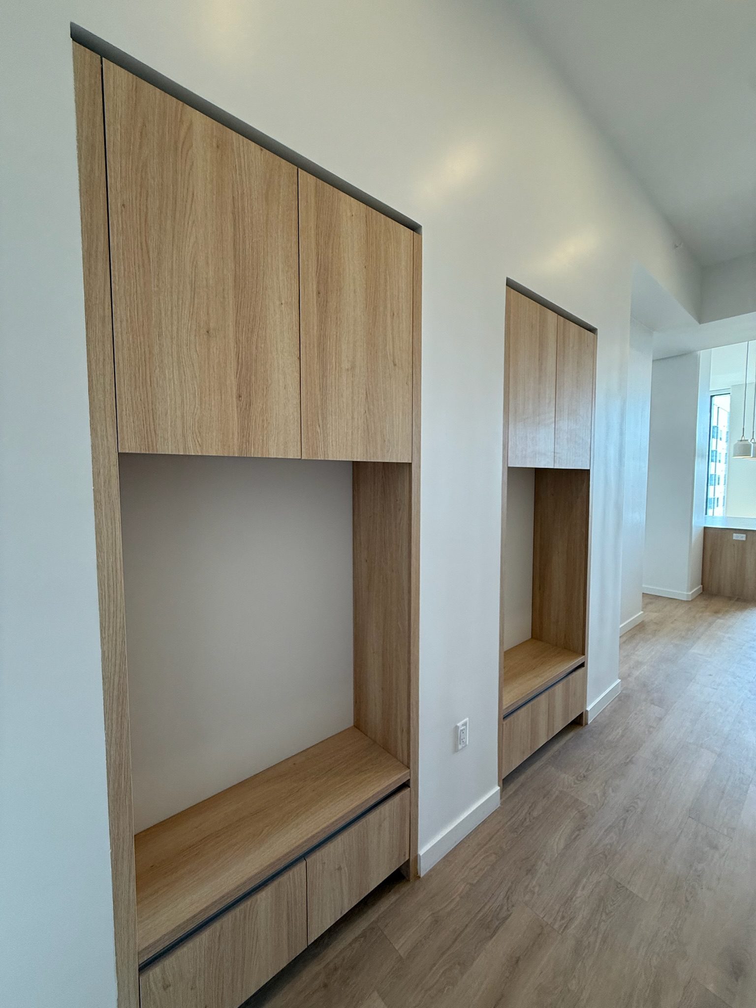 Two built-in wooden alcoves with cabinets above and below, set against a white wall in a modern room with wood flooring and large windows in the background.