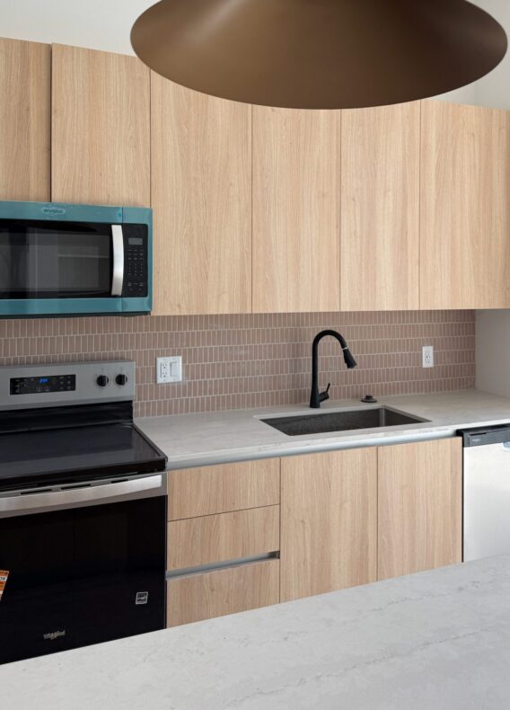 Modern kitchen with light wood cabinets, black stove, microwave, dishwasher, and black faucet. Beige tile backsplash and a bronze pendant light enhance the space. Fresh construction update includes a sleek white countertop.