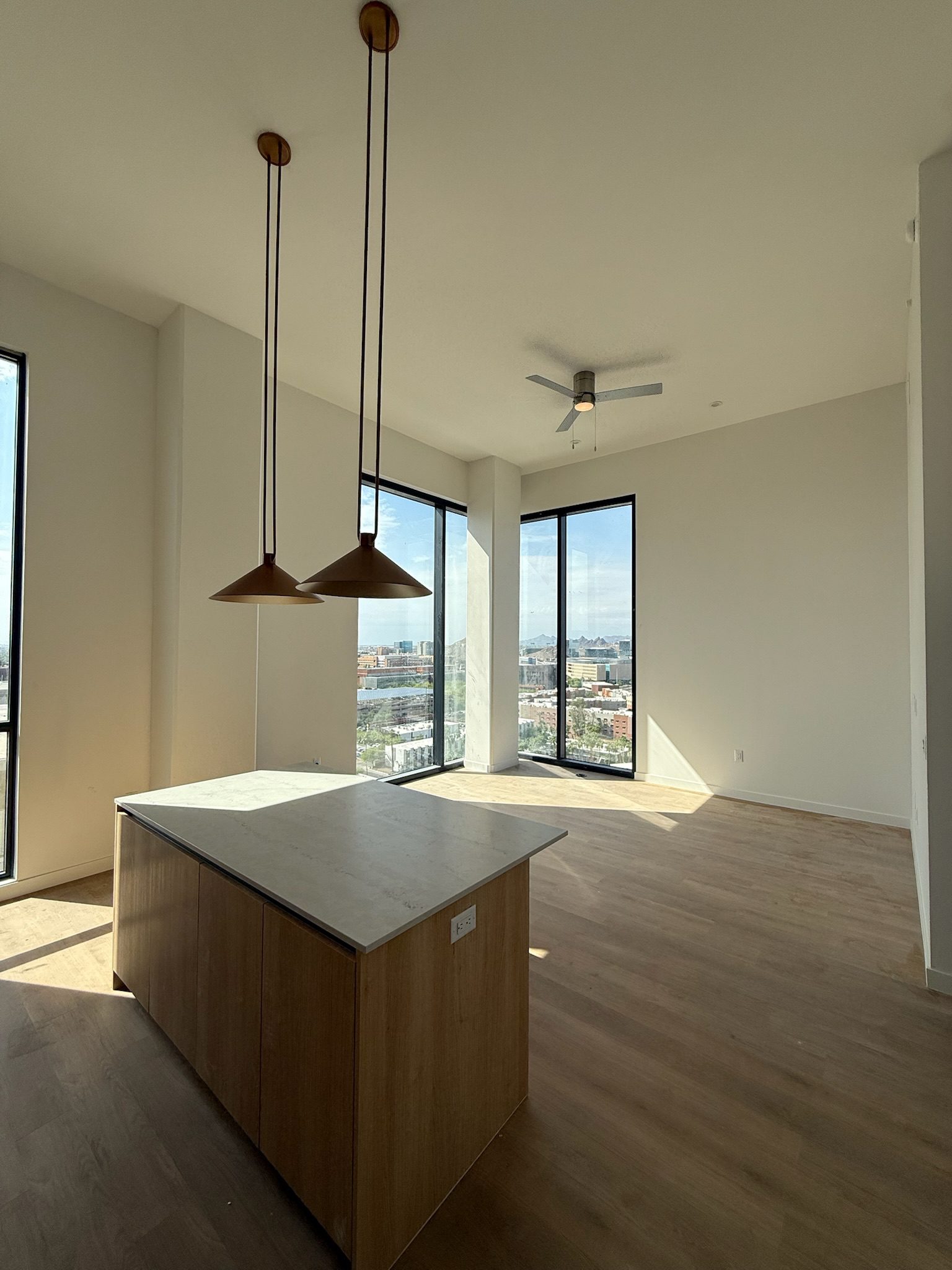 A modern, empty apartment room with large floor-to-ceiling windows, wood floors, a ceiling fan, and an island with two pendant lights above it. Sunlight streams in, offering a view of the city outside.