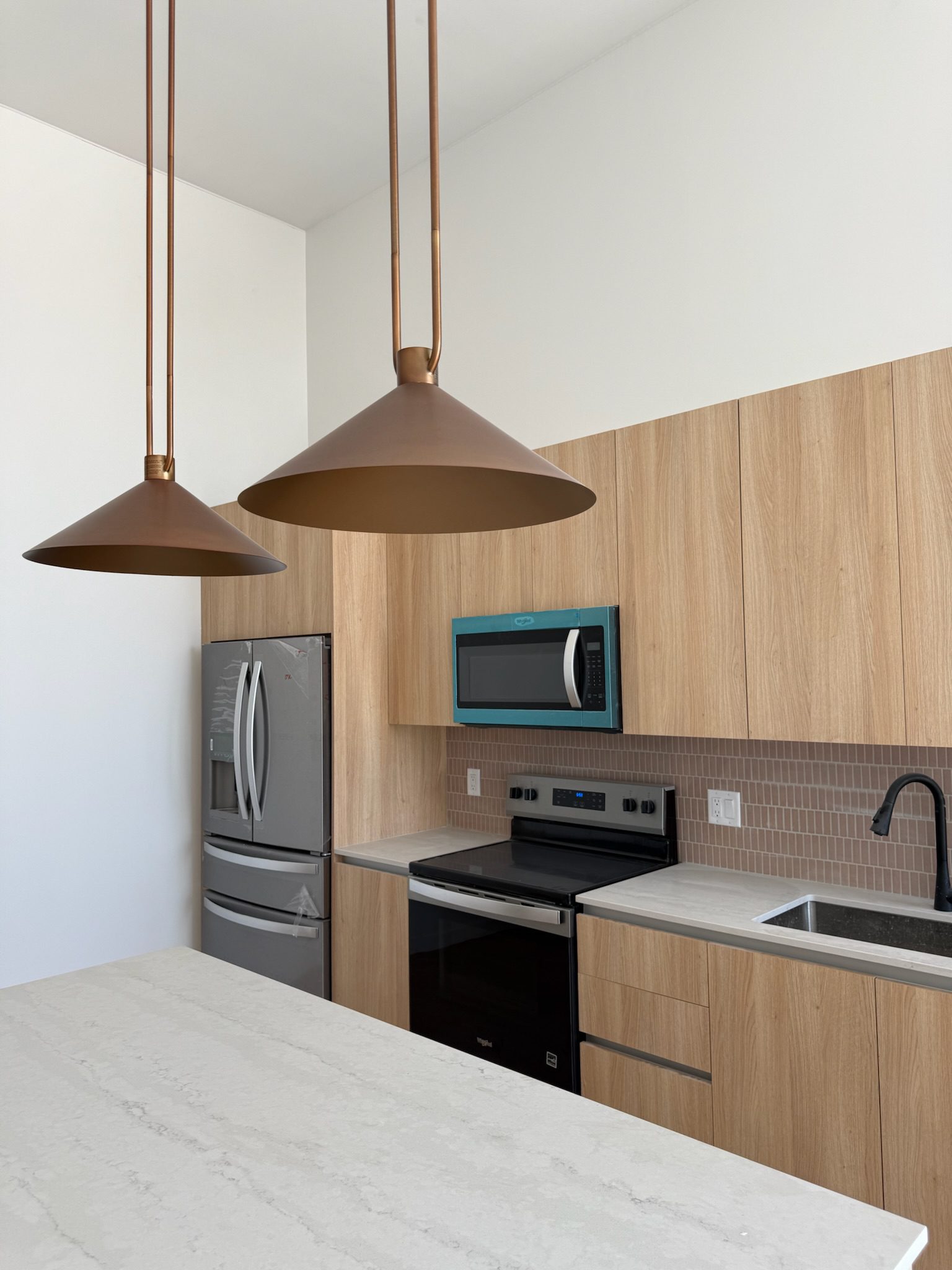 A modern kitchen with light wood cabinets, stainless steel appliances, a built-in microwave, a black faucet, and two large copper pendant lights hanging above a white island countertop.