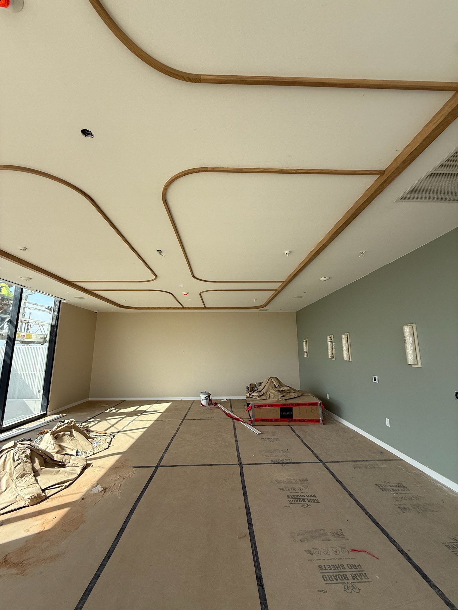 A modern, unfinished room with large windows, cardboard covering the floor, a partially assembled table, and wooden slats forming patterns on the ceiling. Light streams in from the left, highlighting a green accent wall.