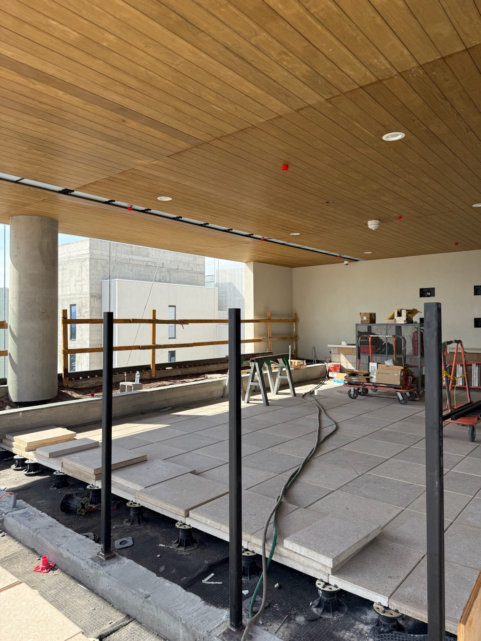 A construction site shows large square tiles being installed on a raised outdoor terrace with metal rail posts, wooden ceiling panels above, and construction tools and materials scattered around.