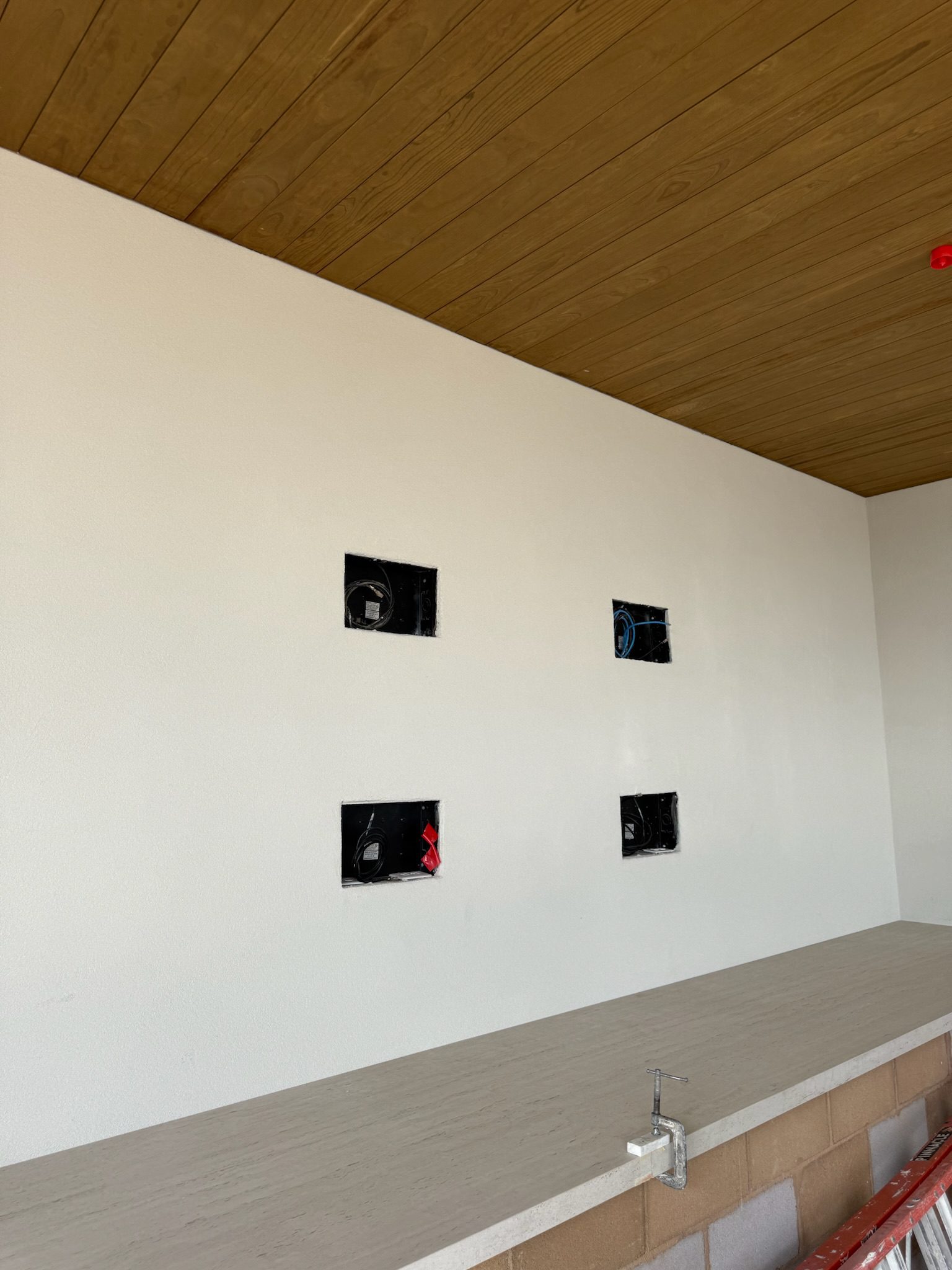 A white wall with four square cutouts exposing electrical wiring, above a countertop with an unfinished faucet. The ceiling is wood-paneled, and the lower wall appears partially constructed.