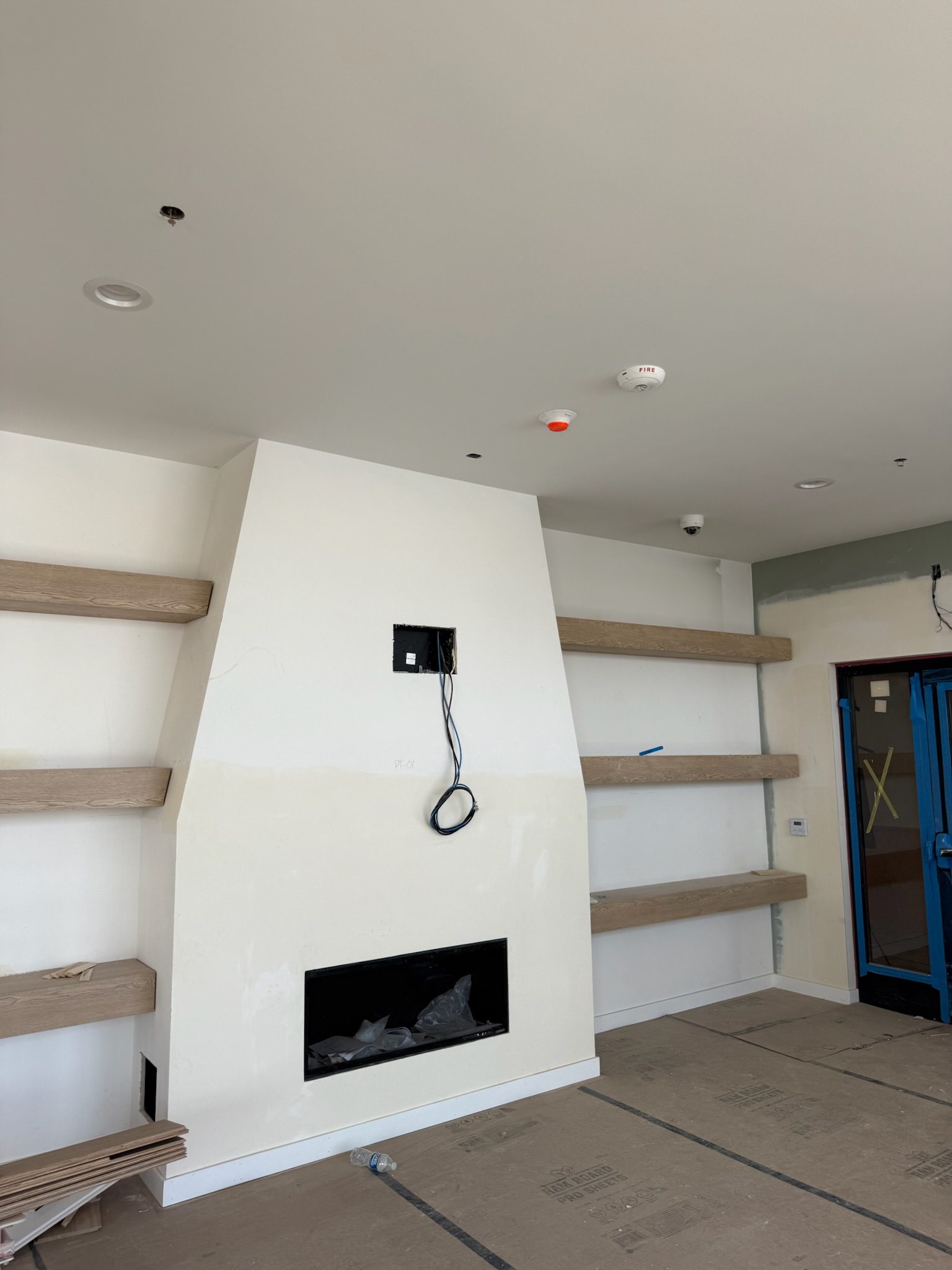 A modern living room under construction with unfinished wooden shelves on both sides of a white fireplace. Electrical wires hang from a cutout above the fireplace. The floor is covered with cardboard.