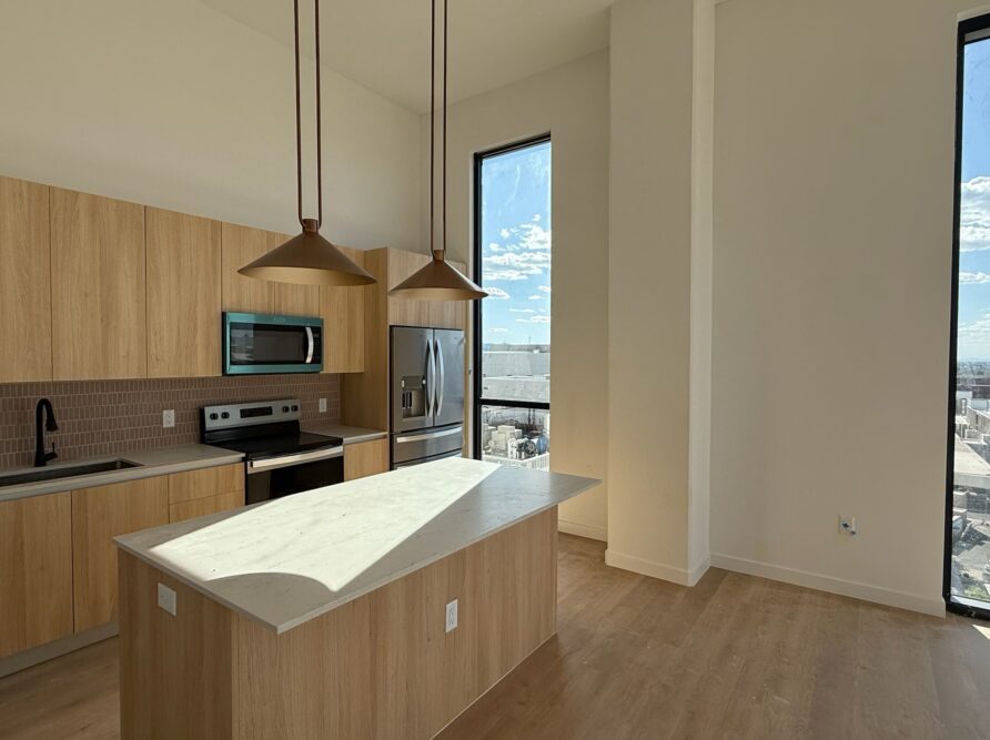 Modern kitchen with light wood cabinets, stainless steel appliances, a center island with white countertop, two pendant lights, and large windows offering a city view under a bright sky—a stunning highlight in the latest construction update.