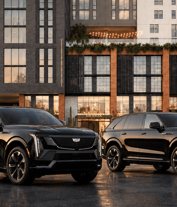 Two black Cadillac SUVs are parked side by side on a wet street in front of a modern, multi-story building with large windows and palm trees, under a partly cloudy sky at sunset.