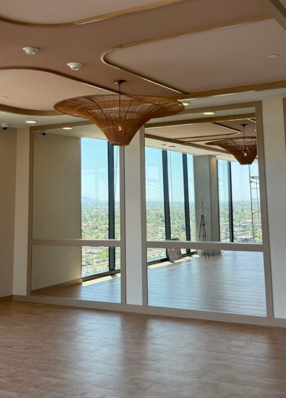 A modern, empty room with large windows and mirrors, tan flooring, and decorative ceiling lights. Sunlight streams in, offering a cityscape view through the glass walls—a recent construction update highlights its sleek design.