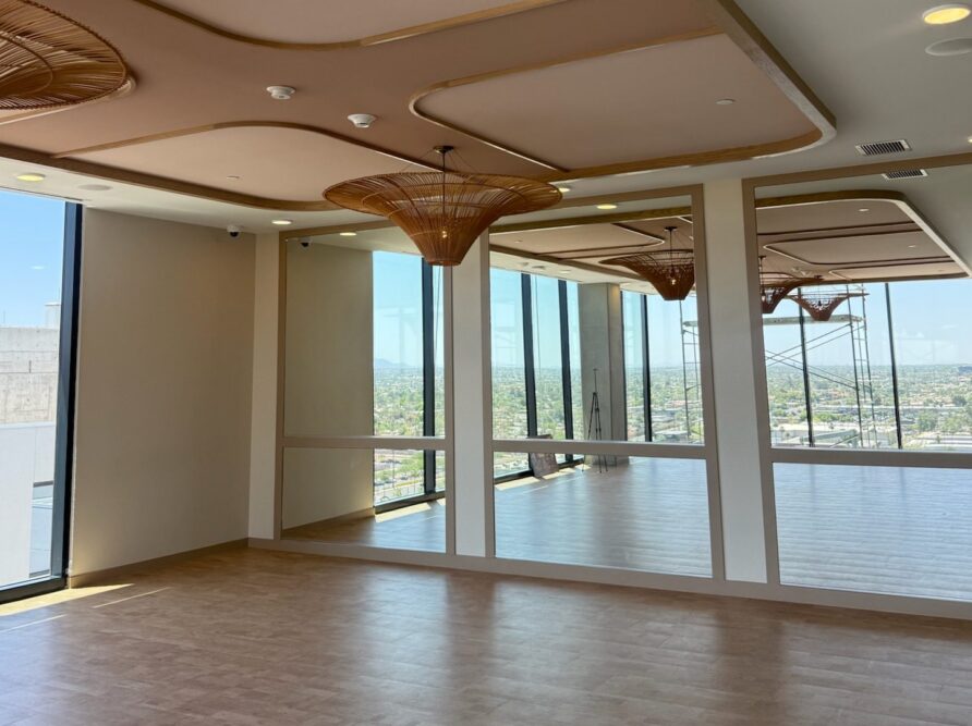 A modern, empty room with large windows and mirrors, tan flooring, and decorative ceiling lights. Sunlight streams in, offering a cityscape view through the glass walls—a recent construction update highlights its sleek design.