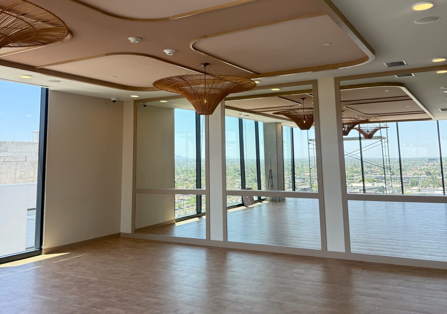 A modern, empty room with large windows and mirrors, tan flooring, and decorative ceiling lights. Sunlight streams in, offering a cityscape view through the glass walls—a recent construction update highlights its sleek design.