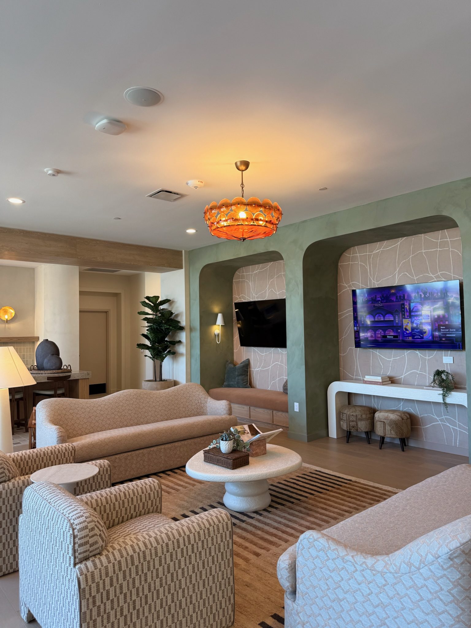 A modern lounge with beige sofas and chairs, a round coffee table, a TV mounted on a green accent wall, decorative shelves, a patterned rug, houseplants, and an orange chandelier. Warm lighting fills the space.