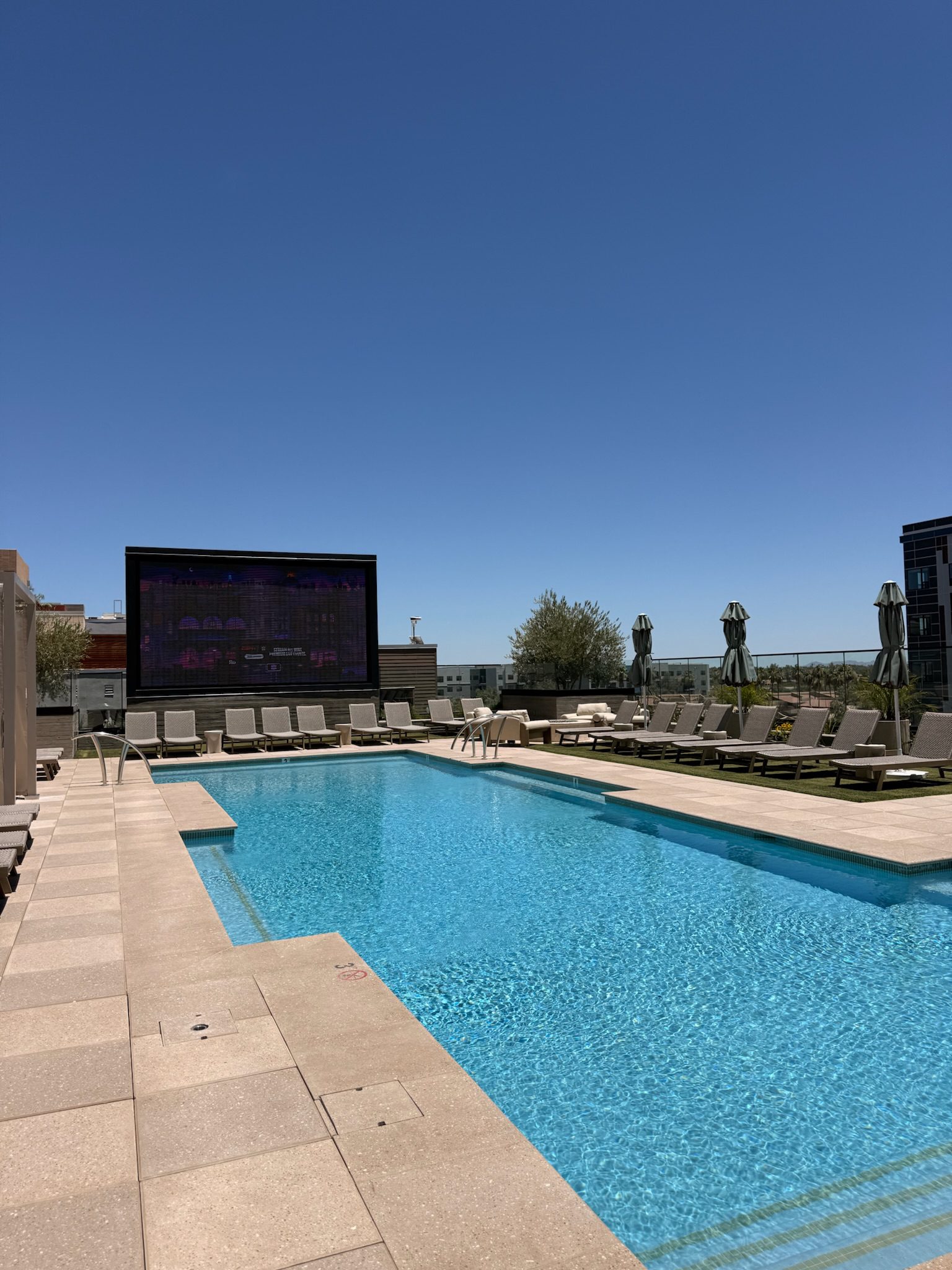 A rooftop swimming pool with lounge chairs and umbrellas beside it, under a clear blue sky. A large screen faces the pool, and city buildings are visible in the background.