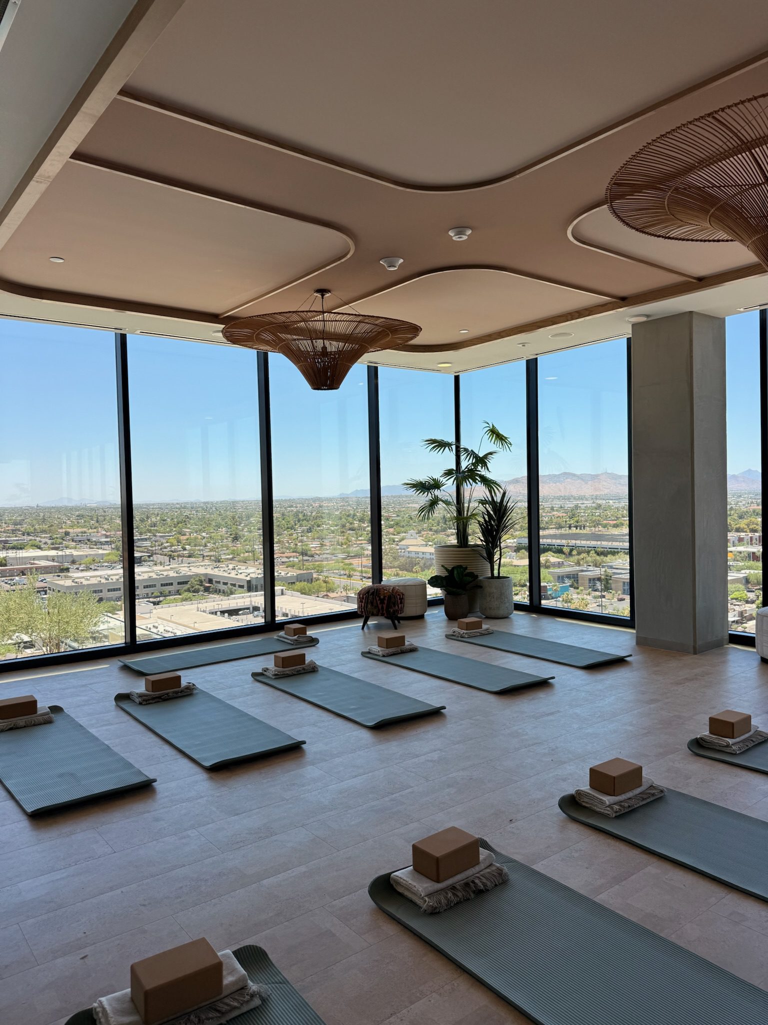 A modern yoga studio with floor-to-ceiling windows, offering a view of a city and distant mountains. Yoga mats, blocks, and towels are neatly arranged on the floor, and there are plants and stylish ceiling lights.