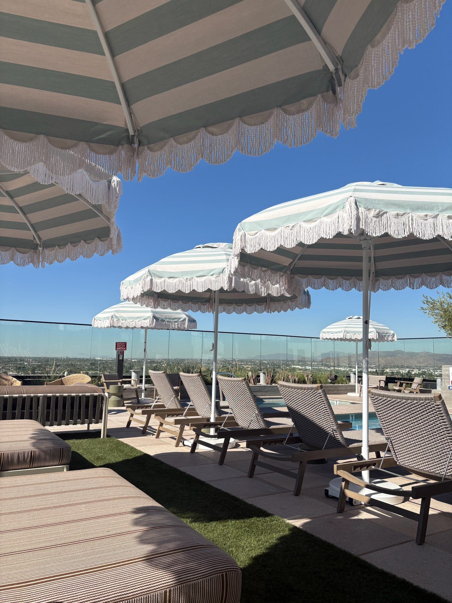 Poolside lounge area with striped umbrellas and empty lounge chairs, set under a clear blue sky. Glass barriers surround the space, offering a view of the landscape beyond.