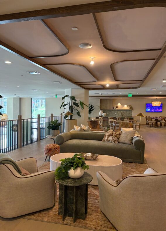 A modern, open lounge area with light-toned sofas and armchairs around a coffee table, plants, and decorative accents. As part of the latest construction update, a TV is mounted above a wooden dining area. Large windows let in natural light.