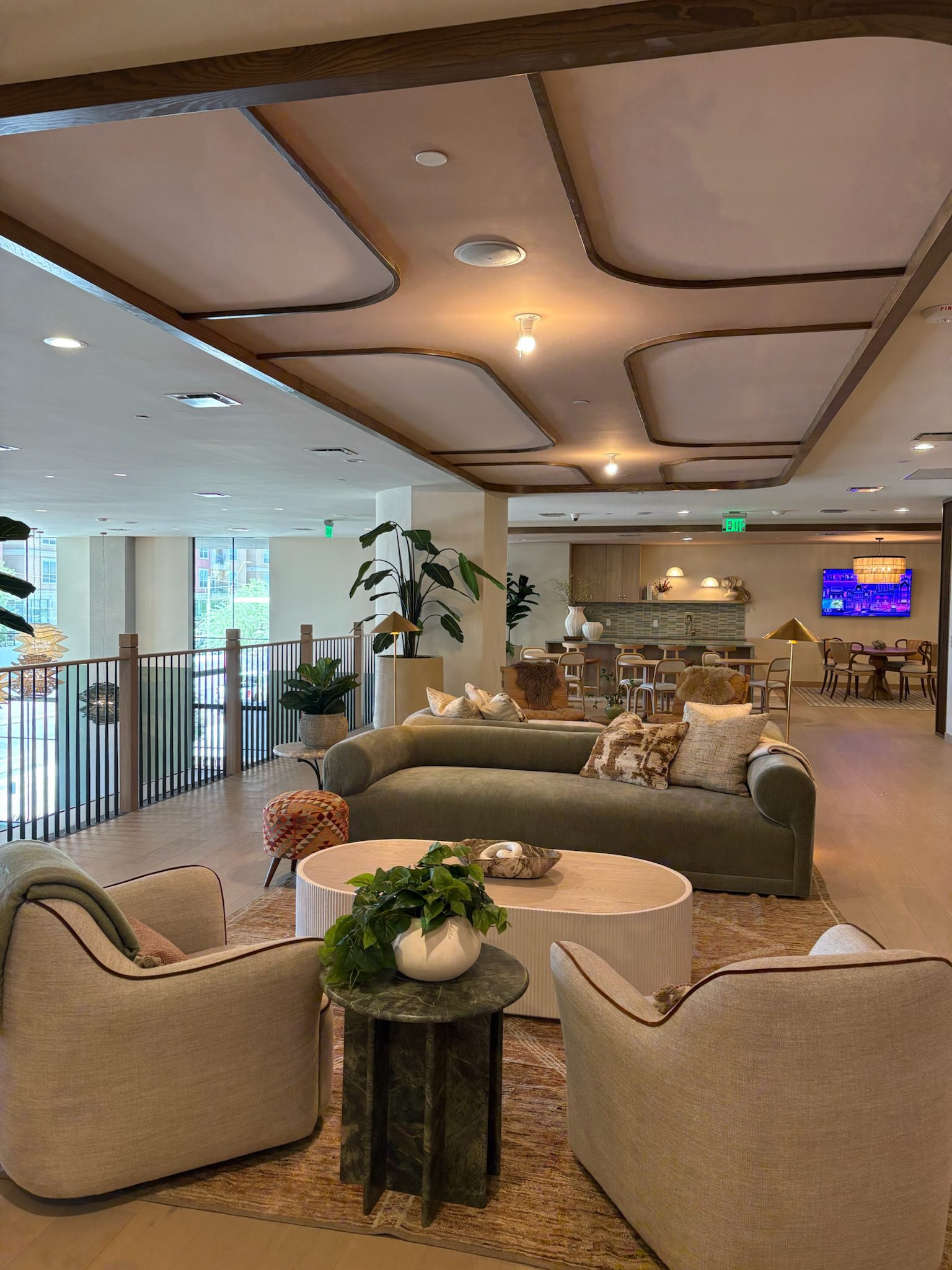 A modern, open lounge area with light-toned sofas and armchairs around a coffee table, plants, and decorative accents. As part of the latest construction update, a TV is mounted above a wooden dining area. Large windows let in natural light.